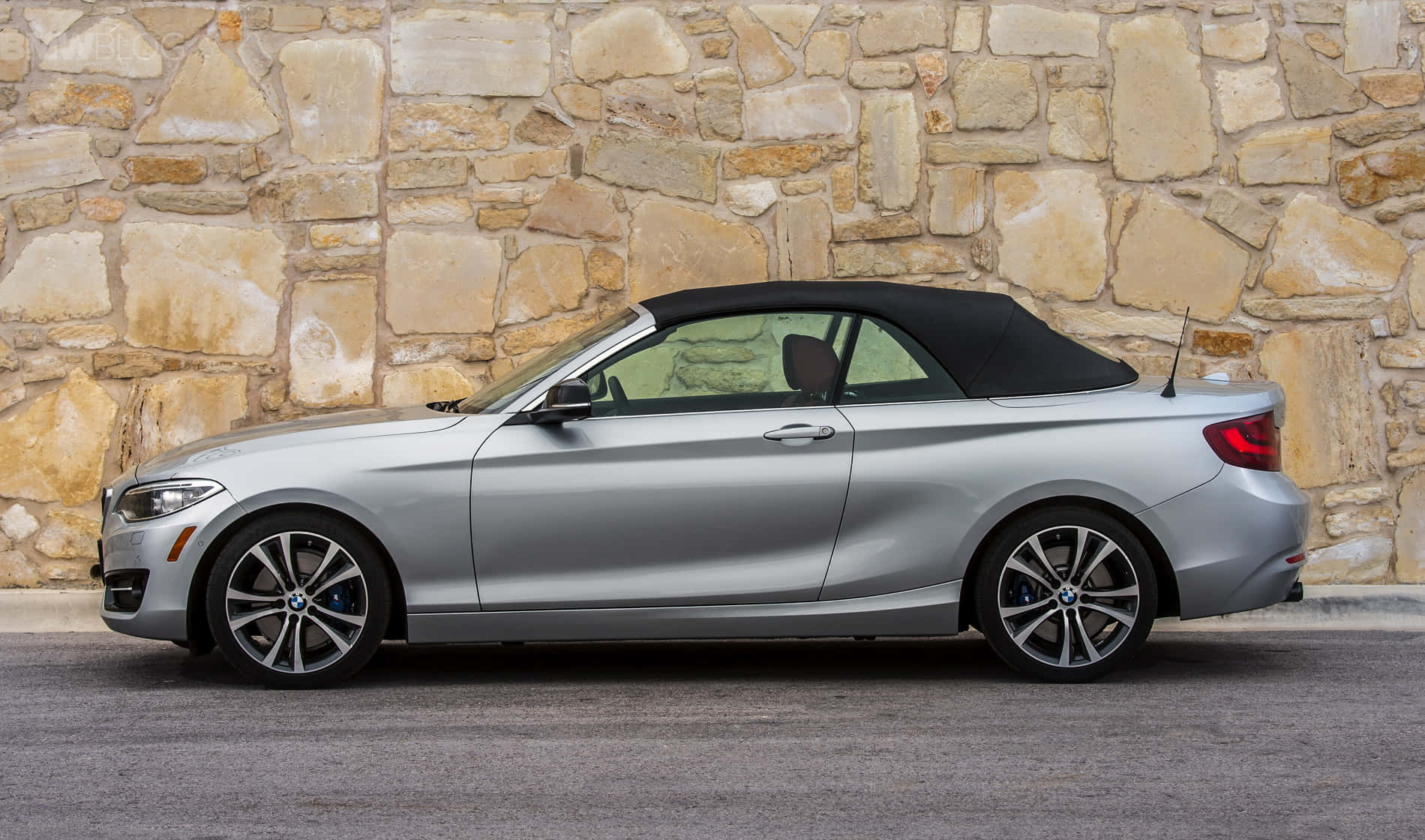 Bmw 2 Series 桌布 壁紙 — 一輛時尚的銀色寶馬敞篷車，配黑色軟頂，停放在質樸的石牆旁。