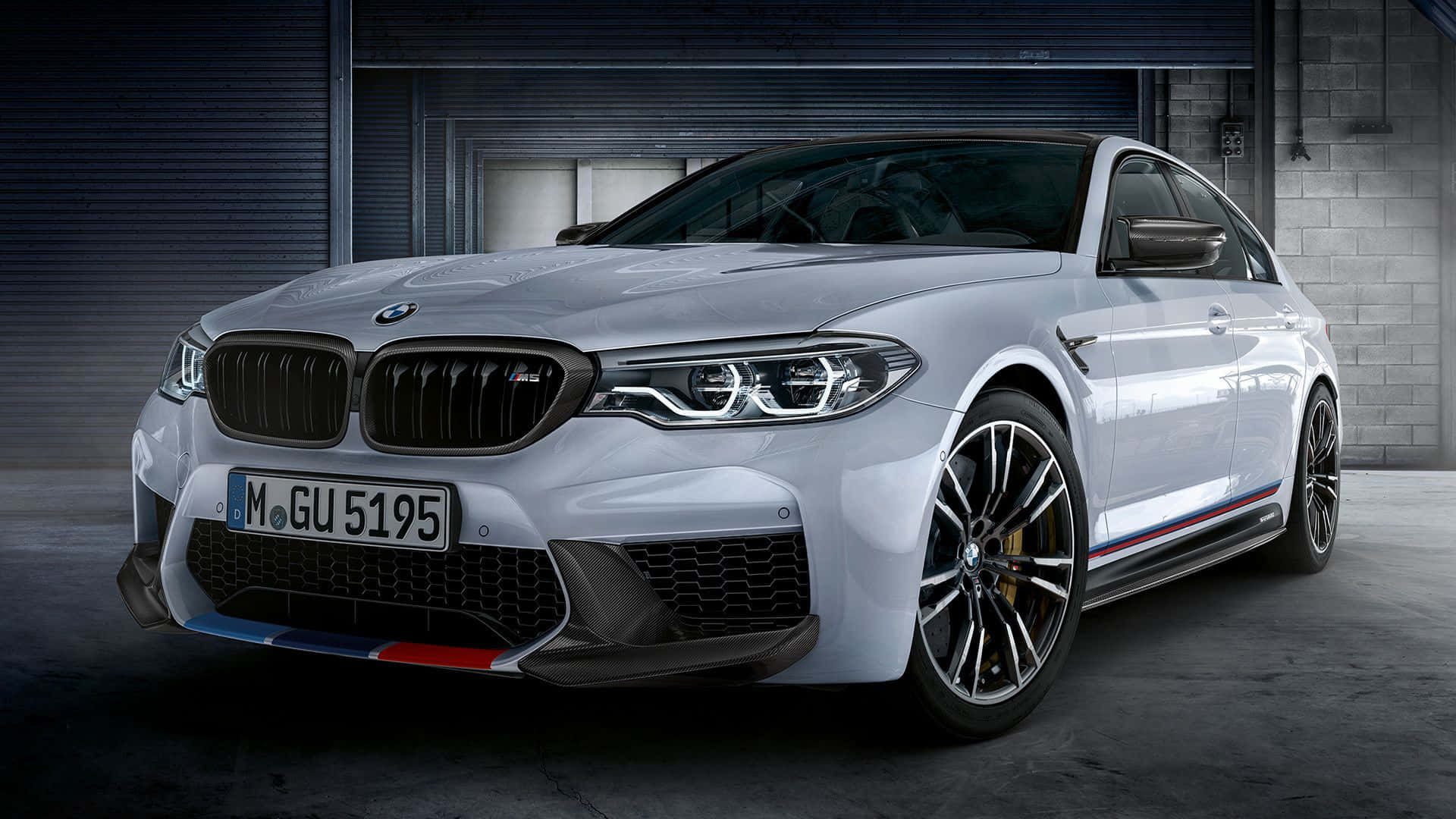 Bmw M5 桌布 壁紙 — 一輛時尚的銀色BMW M5轎車，帶有紅、藍、白賽車條紋，停在光線昏暗的工業車庫中。