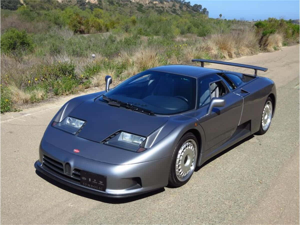 Bugatti Eb110 桌布 壁紙 — 一輛時尚的銀色Bugatti EB110超級跑車停泊在陽光明媚的鄉村道路上，背景有乾草和山丘。