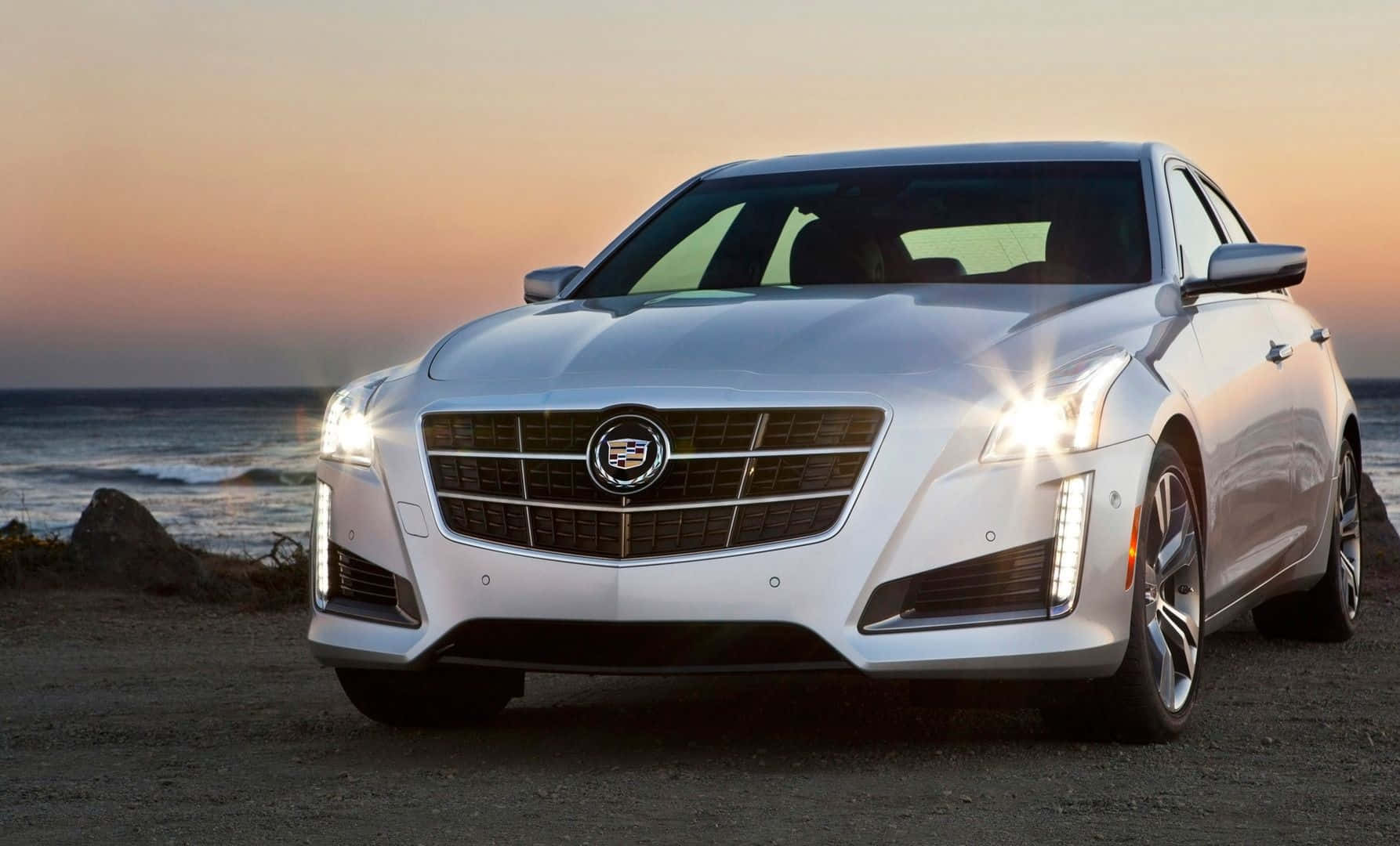 Cadillac Ats 桌布 壁紙 — 一輛時尚的銀色Cadillac CT6停泊在岩石海灘上，夕陽西下，海浪在背後拍打。