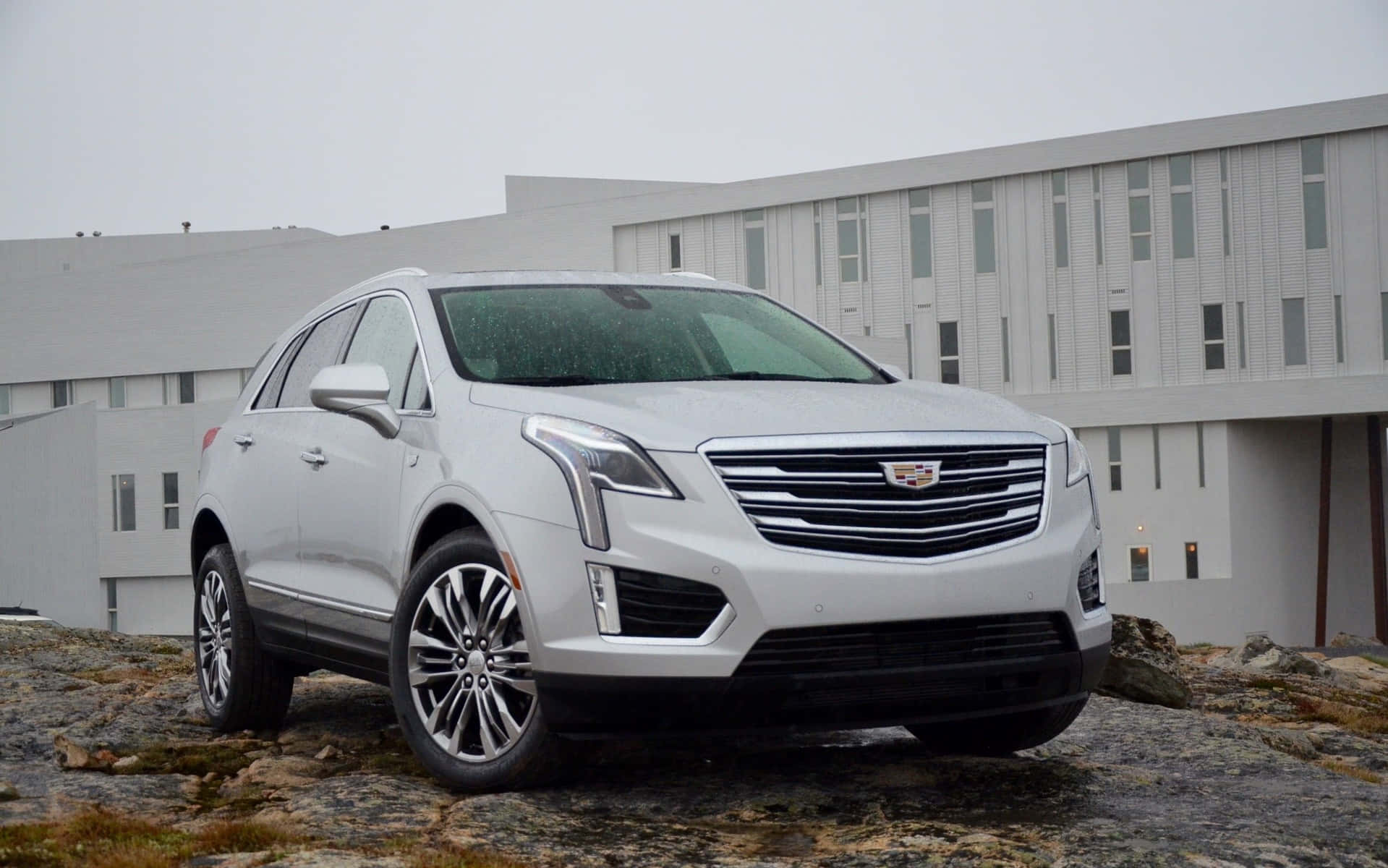 Cadillac Xt5 桌布 壁紙 — 一輛時尚的銀色Cadillac SUV停放在岩石地形上，前方是一座現代灰色建築，天空多雲。