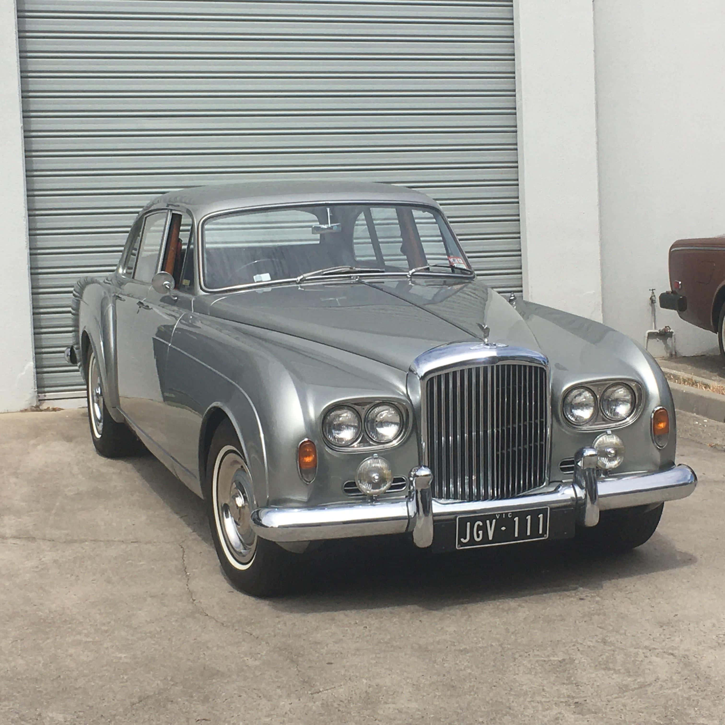 Bentley S3 桌布 壁紙 — 一輛流線型銀色經典賓利轎車停在混凝土車道上的波紋金屬車庫門前。