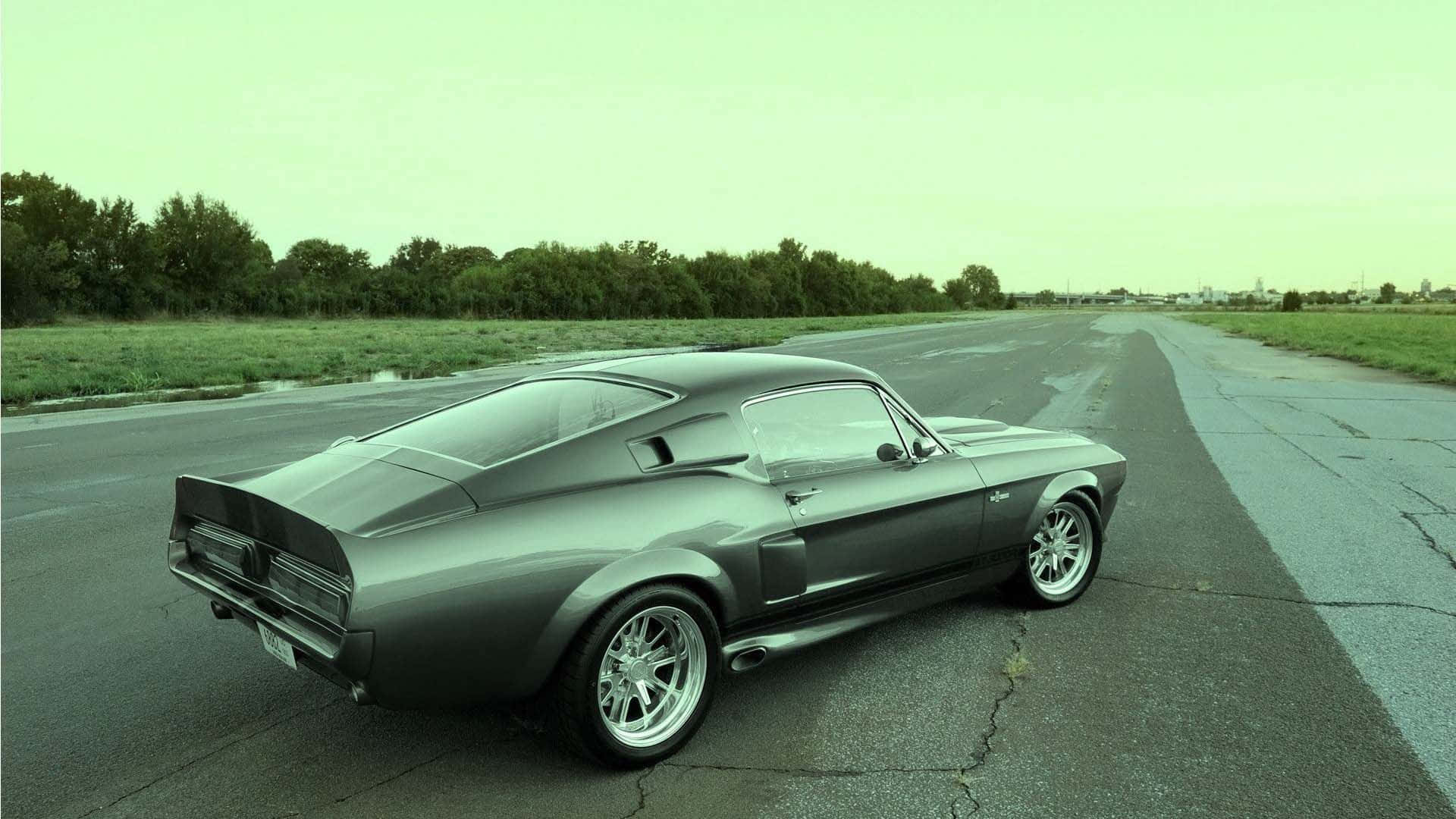Ford Mustang Bullitt 桌布 壁紙 — 一輛時尚的銀色經典肌肉車停在裂開的柏油路上，背景有綠樹和陰天。