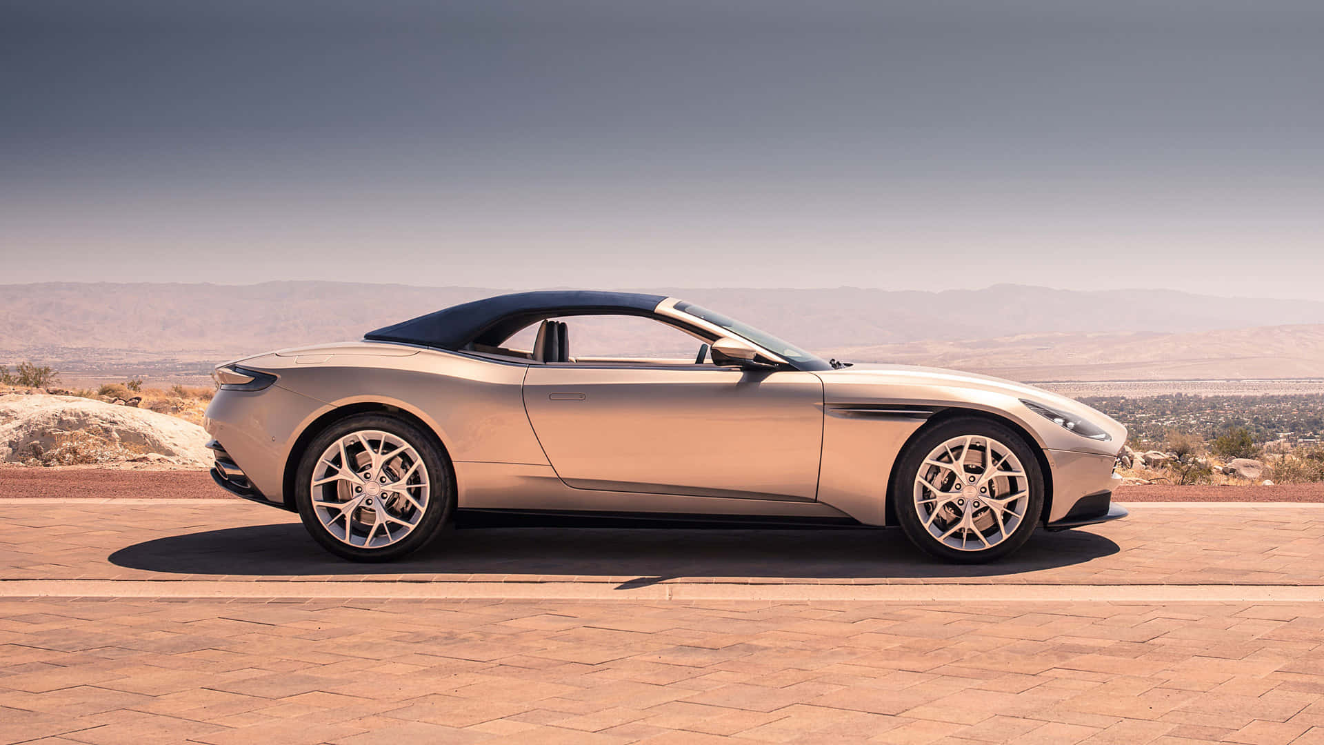Aston Martin Db11 桌布 壁紙 — 一架時尚的銀色敞篷跑車停泊在陽光普照的石頭觀景台上，遠眺朦朧天空下的沙漠山脈。
