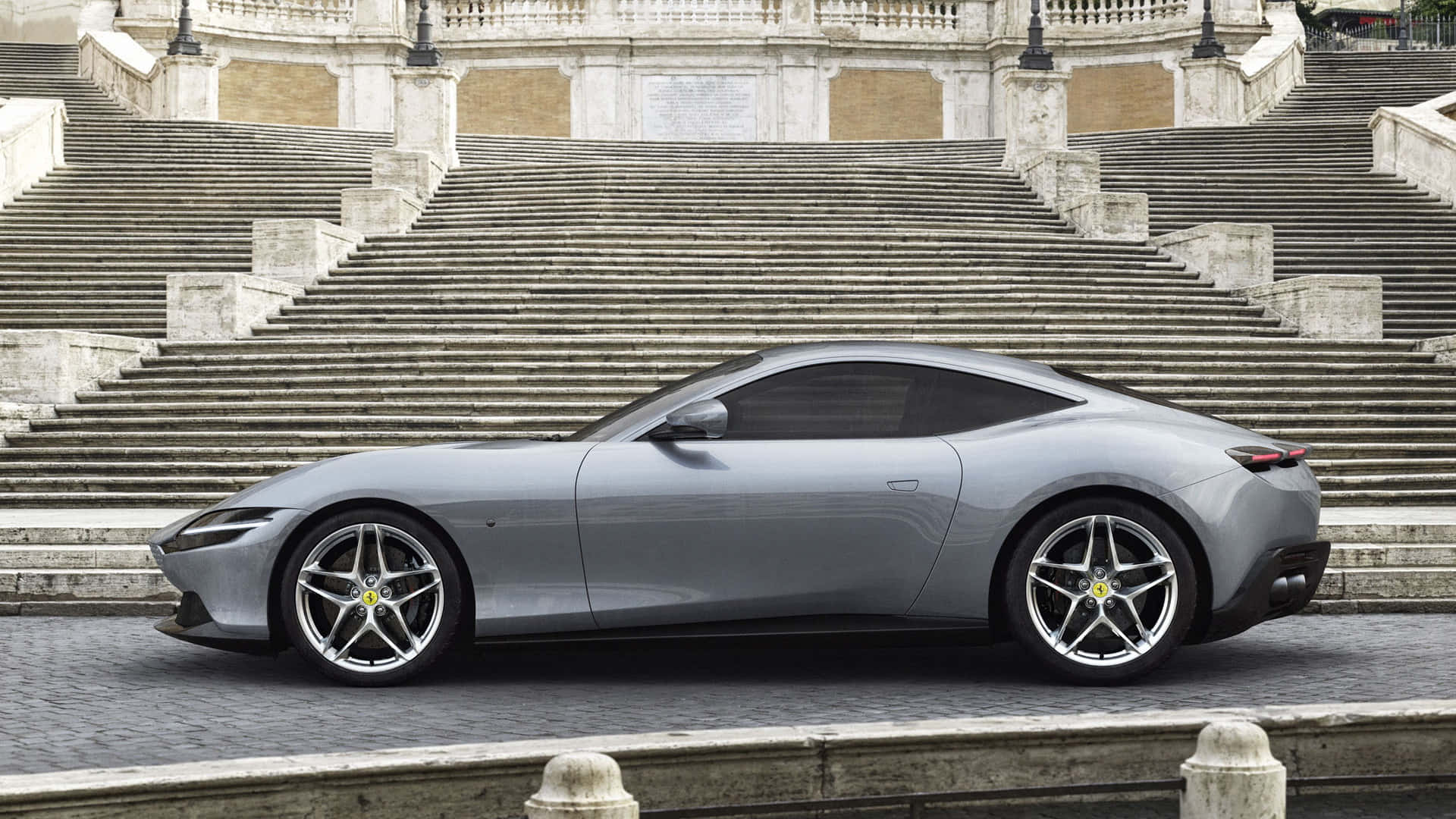 Ferrari Roma 桌布 壁紙 — 一輛時尚的銀色法拉利跑車停放在陽光普照的城市廣場宏偉石階的底部。