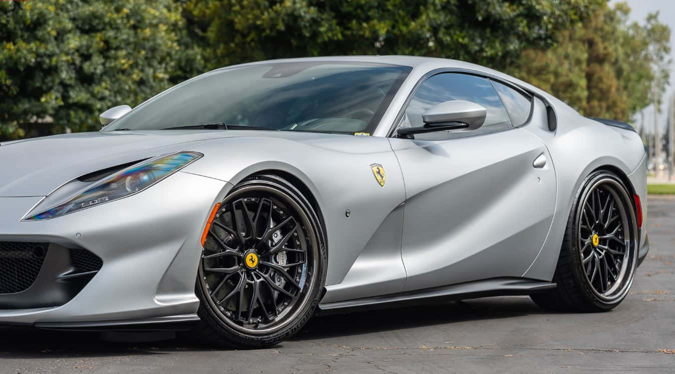Ferrari 812 Superfast 桌布 壁紙 — 一輛時尚的銀色法拉利跑車，配備黑色車輪，在明亮的日光下停泊在路面上，背景有綠樹。