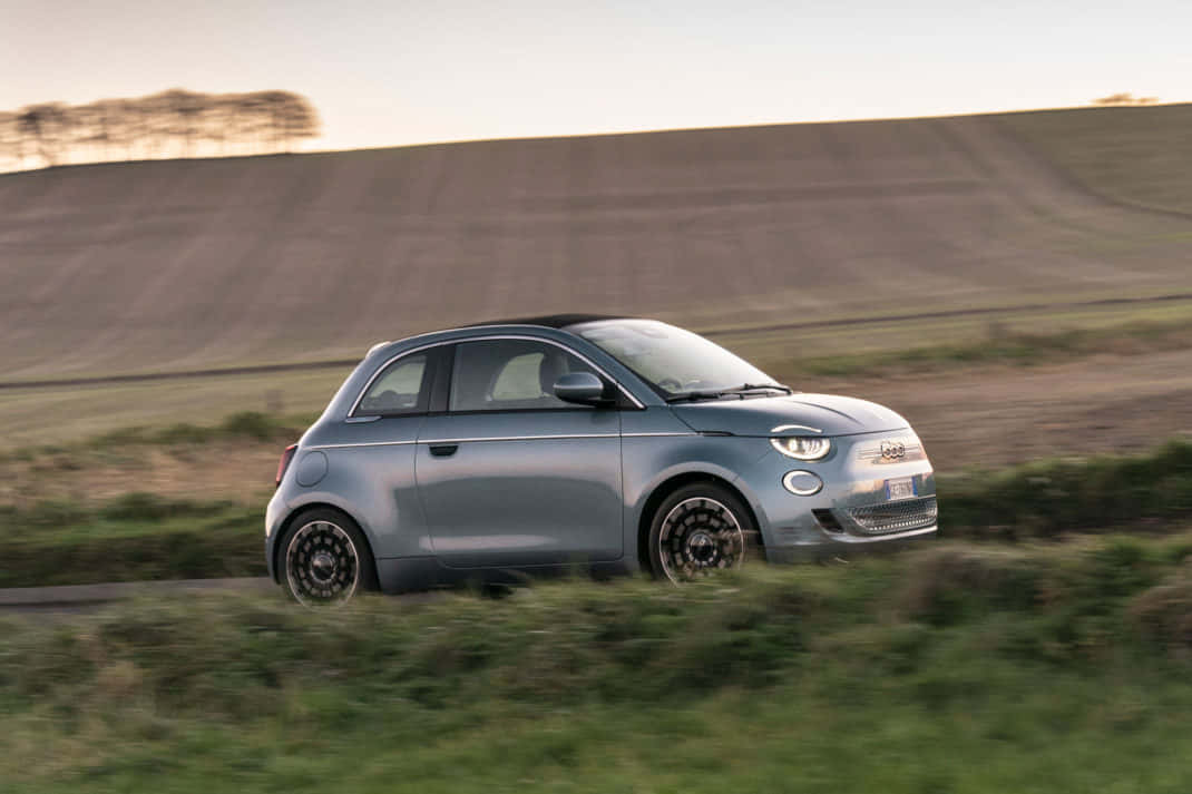 Fiat 500 桌布 壁紙 — 一輛時尚的銀色Fiat 500e電動車在黃金時段沿鄉村道路行駛，背景是模糊的綠色田野和起伏的山丘。