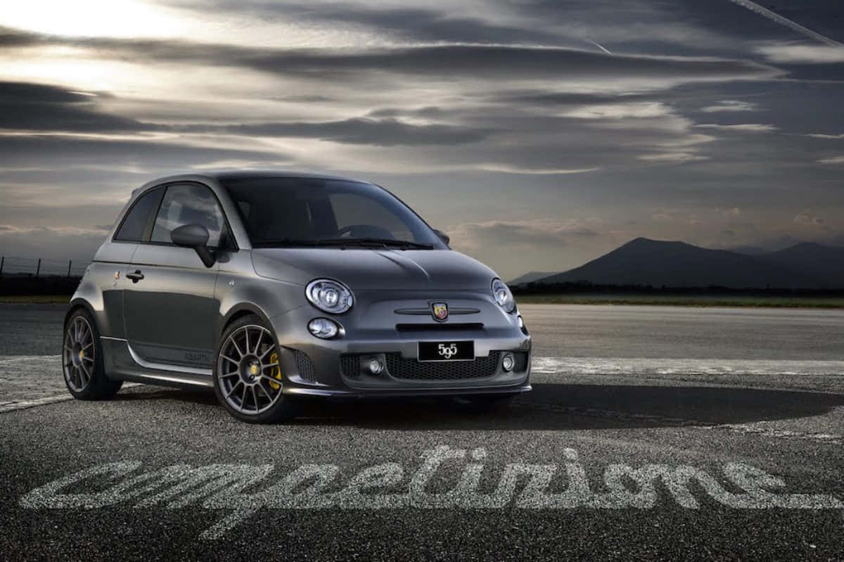 Fiat 500 桌布 壁紙 — 一架時尚的銀色Fiat 599跑車停在瀝青跑道上，背景是戲劇性的多雲天空和遠處的山脈。