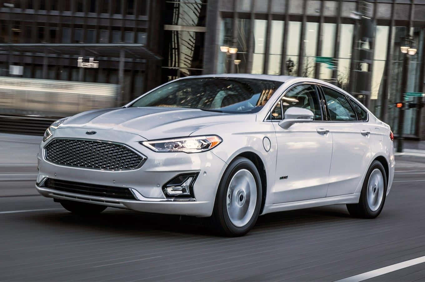 Ford Fusion 桌布 壁紙 — 一輛時尚的銀色Ford Fusion Hybrid在現代玻璃建築背景下，以動態模糊效果行駛在城市街道上。