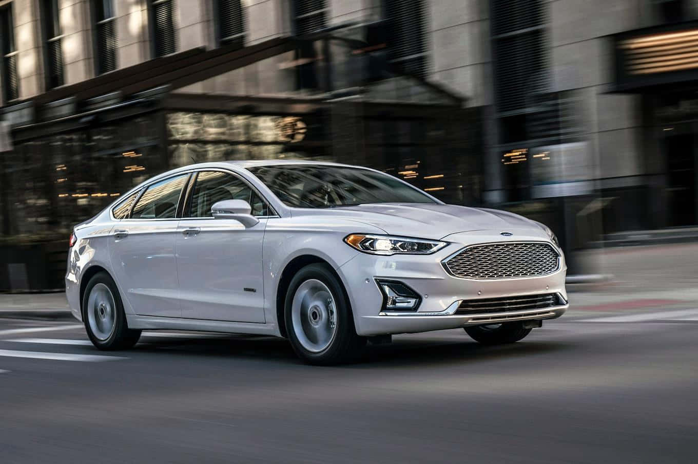 Ford Fusion 桌布 壁紙 — 一輛時尚的銀色福特Fusion轎車在黃昏時分高速駛過模糊的城市街道，背景是現代玻璃建築。
