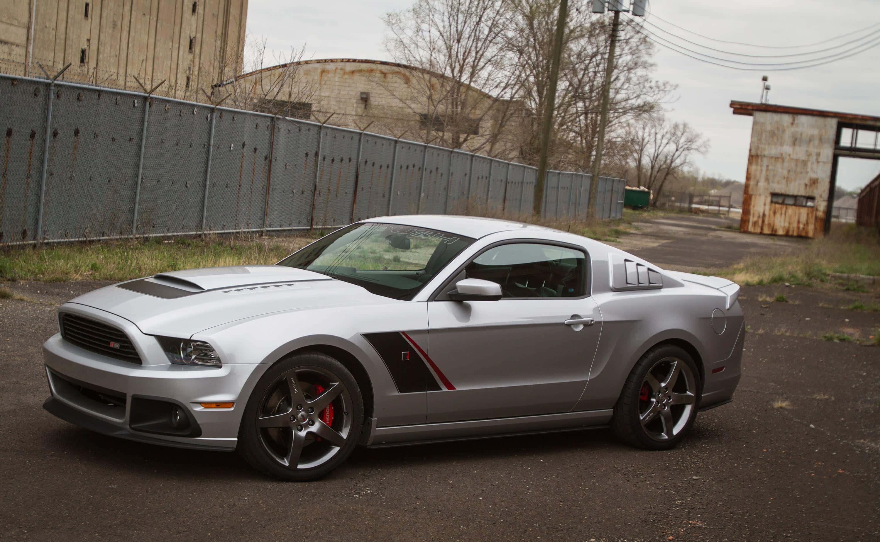 Ford Mustang Roush 桌布 壁紙 — 一架時尚的銀色福特野馬Shelby GT500停放在工業用地，周圍有生鏽的圍欄和倉庫，天空多雲。