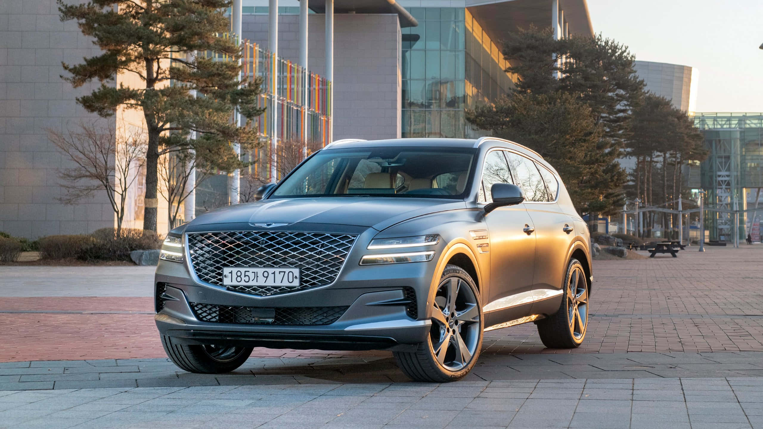 Genesis G70 桌布 壁紙 — 一輛時尚的銀色Genesis GV80 SUV停泊在鋪砌的廣場上，背景是現代玻璃建築和樹木，正值黃金時段。