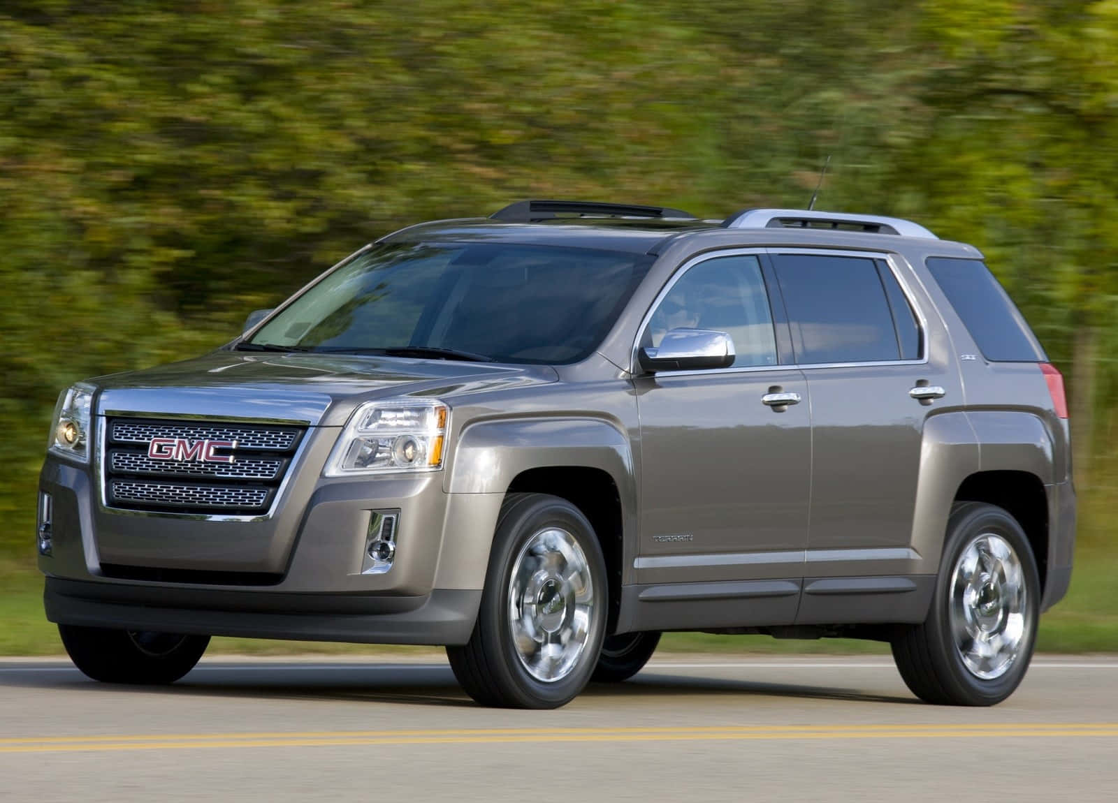 Gmc Terrain 桌布 壁紙 — 一輛時尚的銀色GMC Terrain SUV在陽光明媚的道路上行駛，背景是模糊的綠色樹葉。
