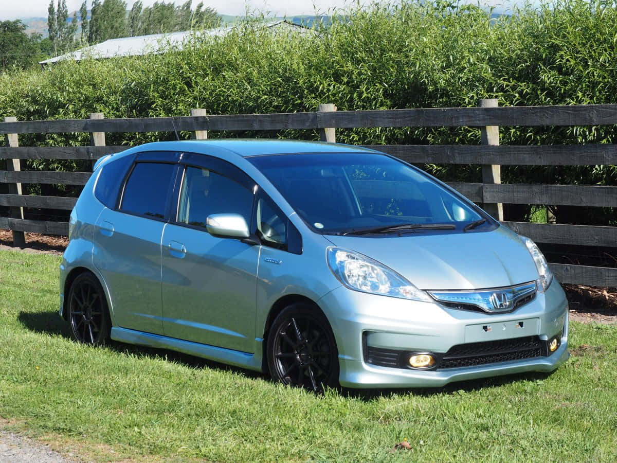 Honda Fit 桌布 壁紙 — 一輛時尚的銀色本田Fit停在草地上，旁邊有木柵欄，背景是茂密的綠色樹籬。