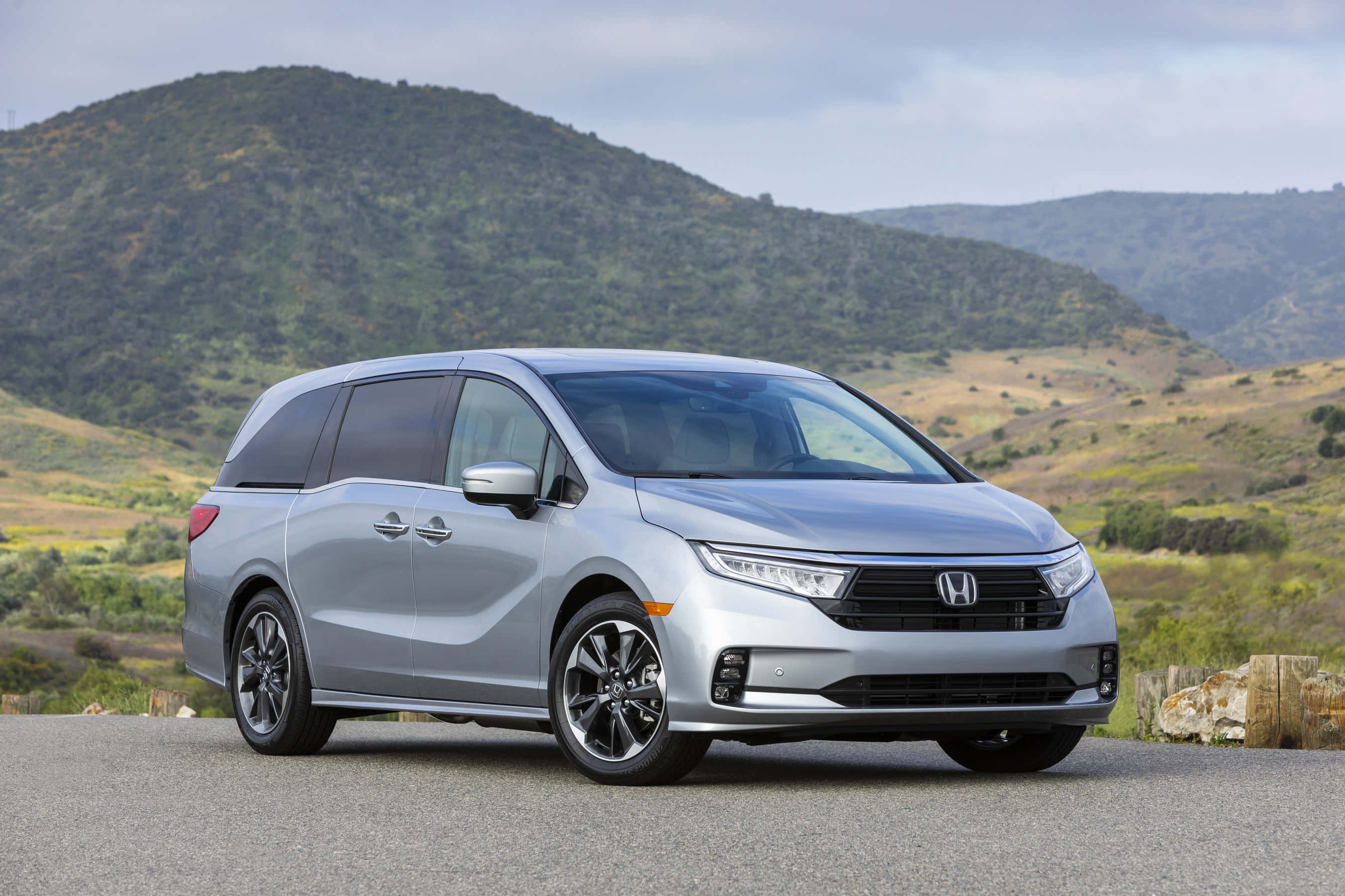 Honda Odyssey 桌布 壁紙 — 一輛流線型銀色本田小型貨車停在鋪砌道路上，背景是起伏的綠色山丘和多雲的天空。