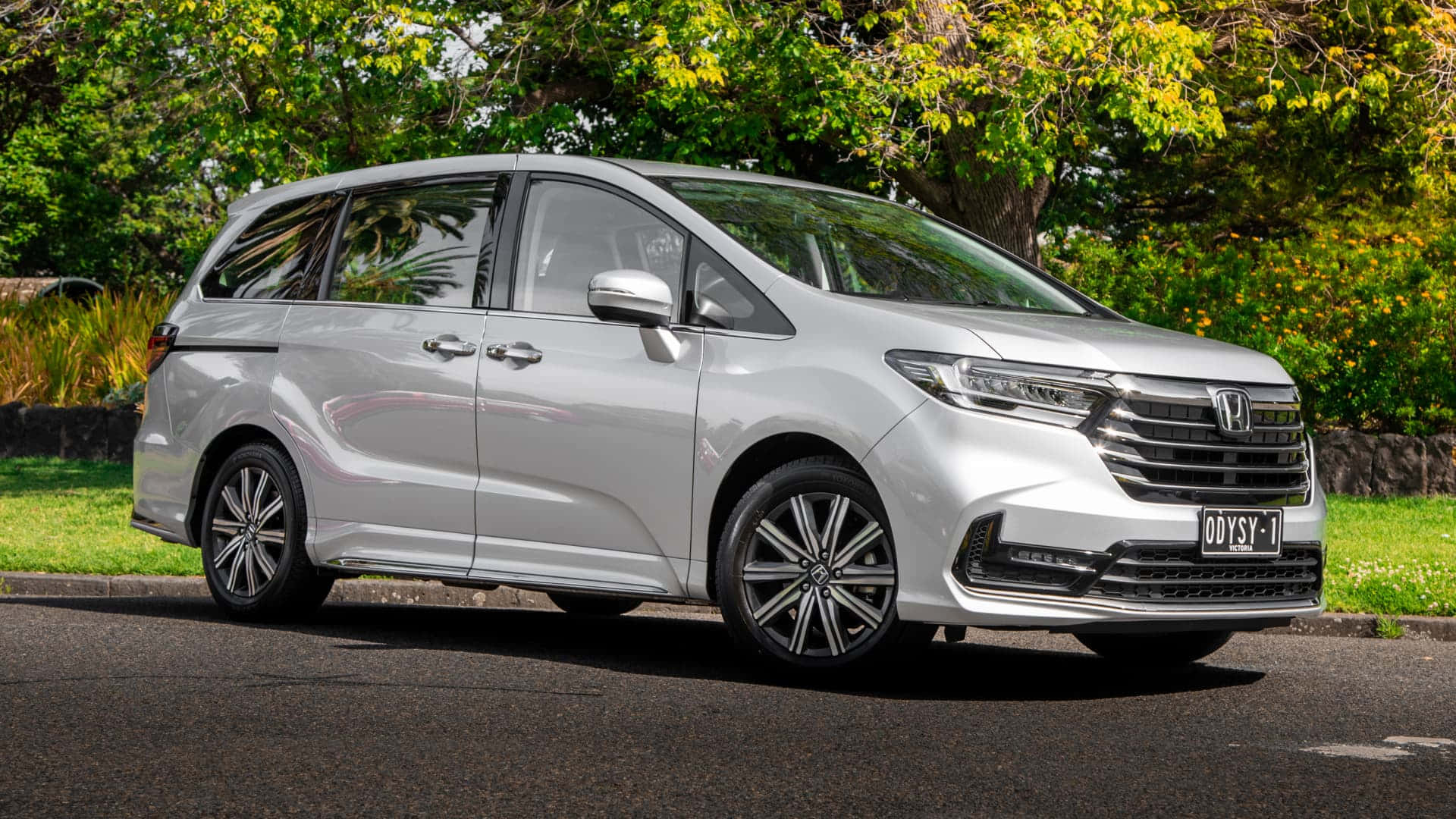 Honda Odyssey 桌布 壁紙 — 一輛流線型銀色本田Odyssey廂型車停放在鋪砌的道路上，背景有茂密的綠樹和黃花。