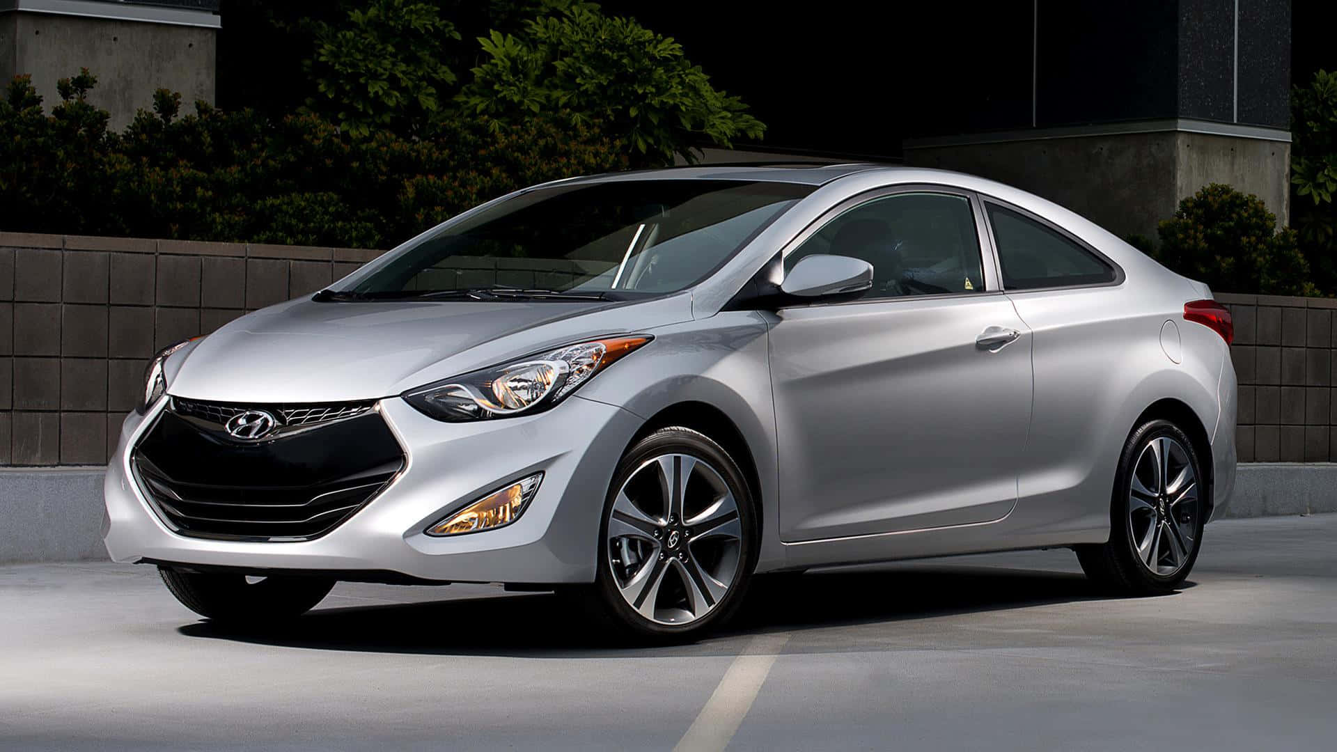 Hyundai Elantra 桌布 壁紙 — 一輛時尚的銀色現代轎跑車停在混凝土表面上，背景有深色樹葉和混凝土牆。
