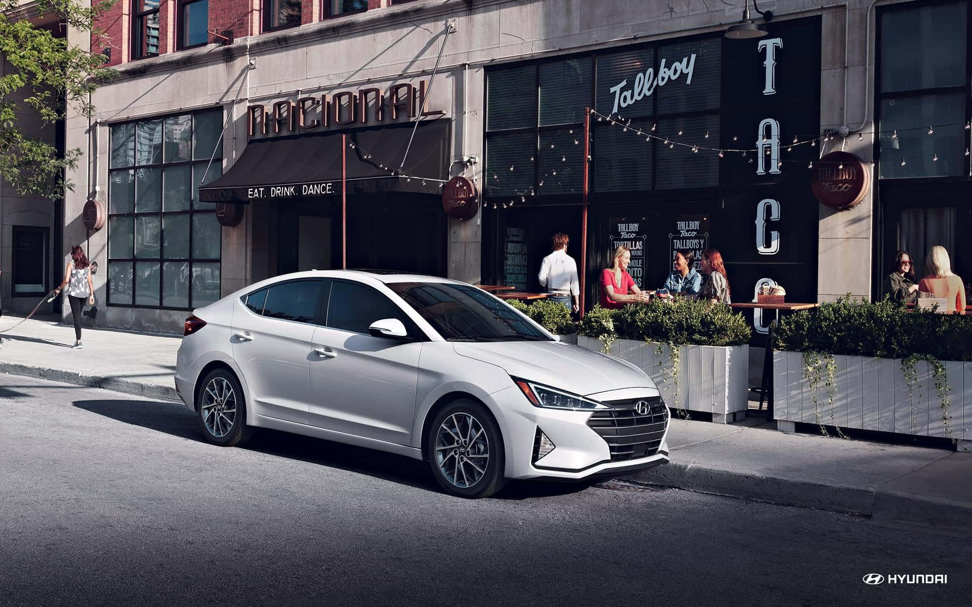 Hyundai Elantra 桌布 壁紙 — 一架時尚的銀色現代Elantra停放在陽光明媚的城市街道上，旁邊是一間設有戶外座位的熱鬧墨西哥捲餅餐廳。