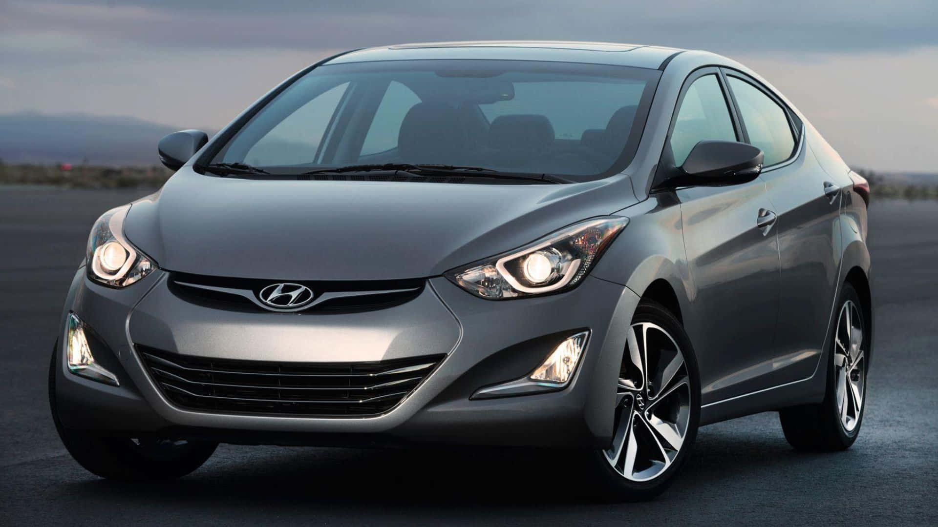 Hyundai Elantra 桌布 壁紙 — 一輛時尚的銀色現代伊蘭特轎車在柔和暮光下，在開闊的瀝青路上閃耀，遠方有山丘。
