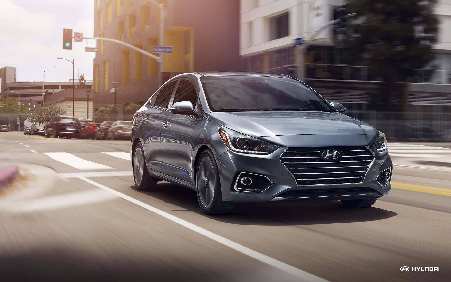 Hyundai Accent 桌布 壁紙 — 一輛時尚的銀色現代轎車在陽光普照的城市街道上飛馳，背景建築物和道路呈現動態模糊效果。