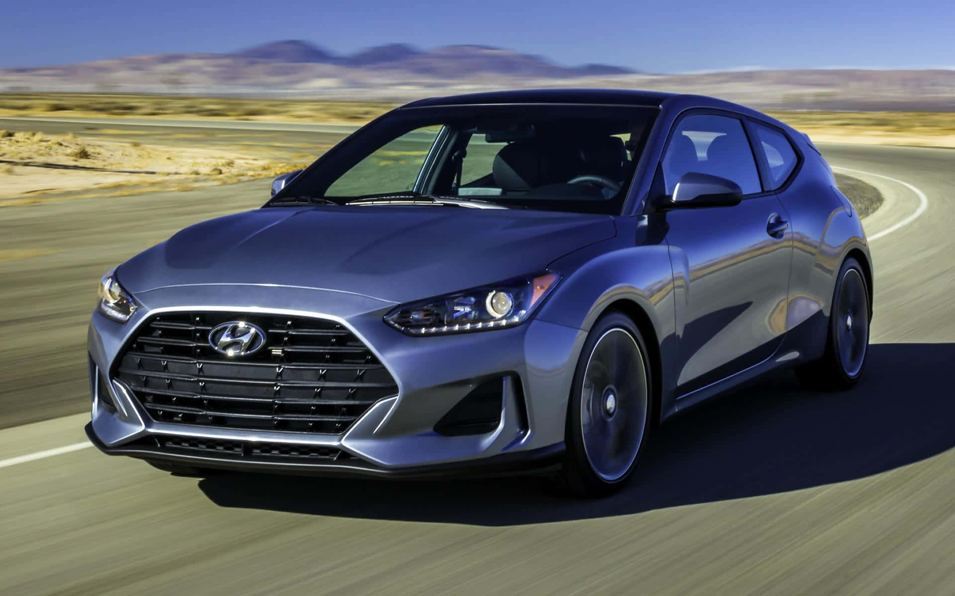 Hyundai Veloster 桌布 壁紙 — 一輛時尚的銀色Hyundai Veloster在晴朗藍天和遠山背景下，沿沙漠高速公路飛馳。