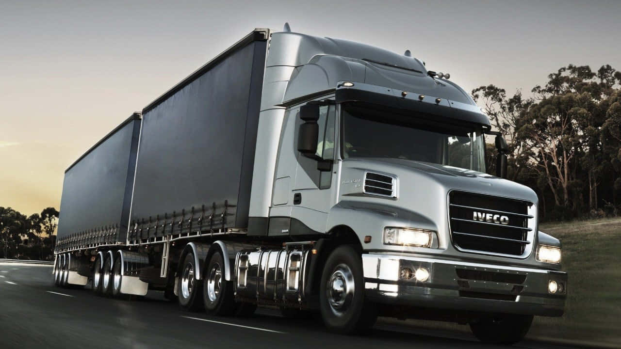  壁紙 — 一輛時尚的銀色 Iveco 半掛卡車拖著黑色拖車，在日落時分行駛在兩旁樹木林立的公路上。