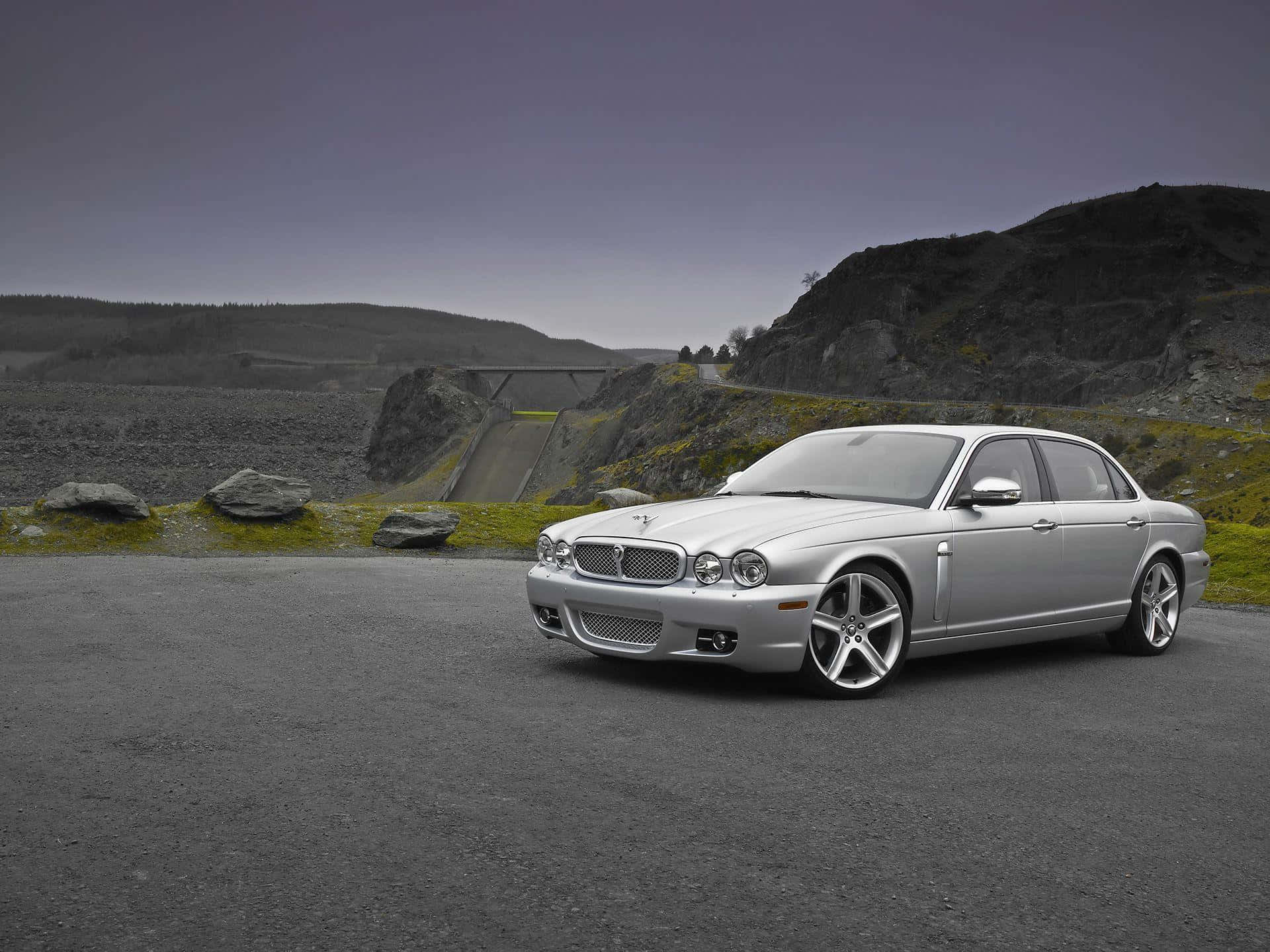 Jaguar Xjr 桌布 壁紙 — 一輛流線型銀色積架轎車停放在鋪砌道路上，旁邊是岩石山坡，天空呈現柔和的灰色。