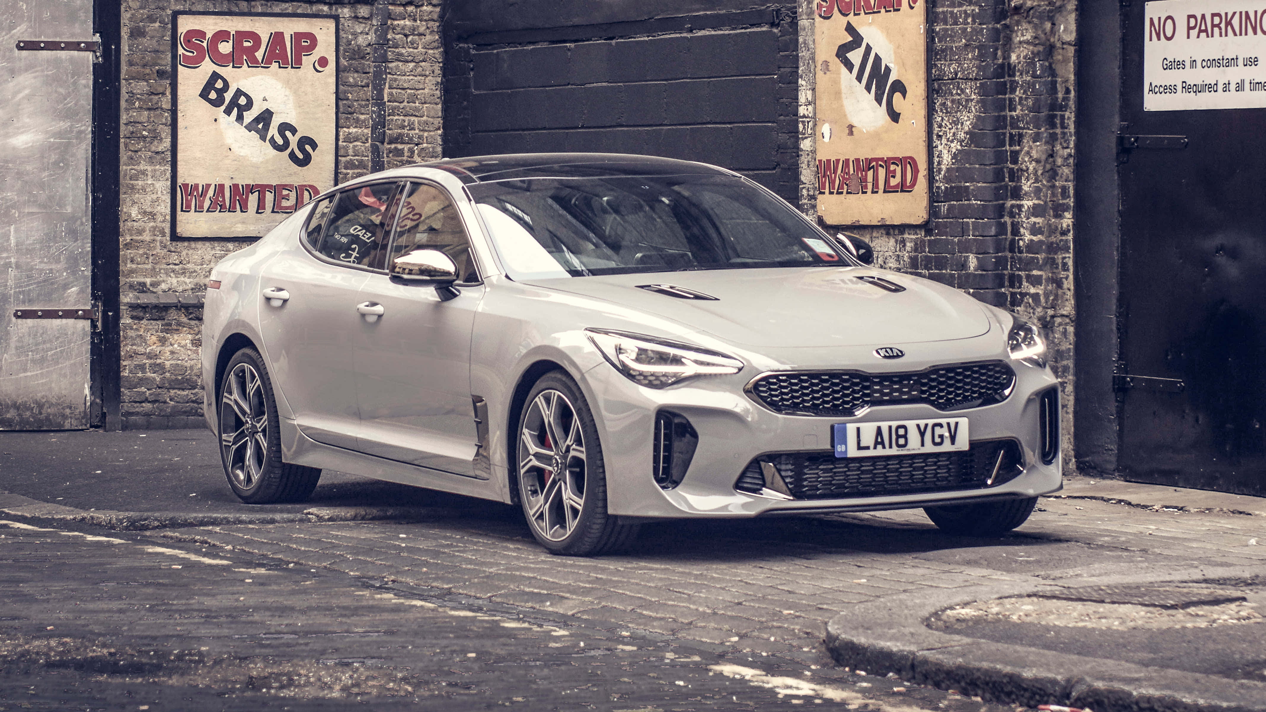 Kia Stinger 桌布 壁紙 — 一台時尚的銀色Kia Stinger運動轎車停泊在鵝卵石街道上，旁邊是帶有復古廢金屬收購標誌的磚牆。