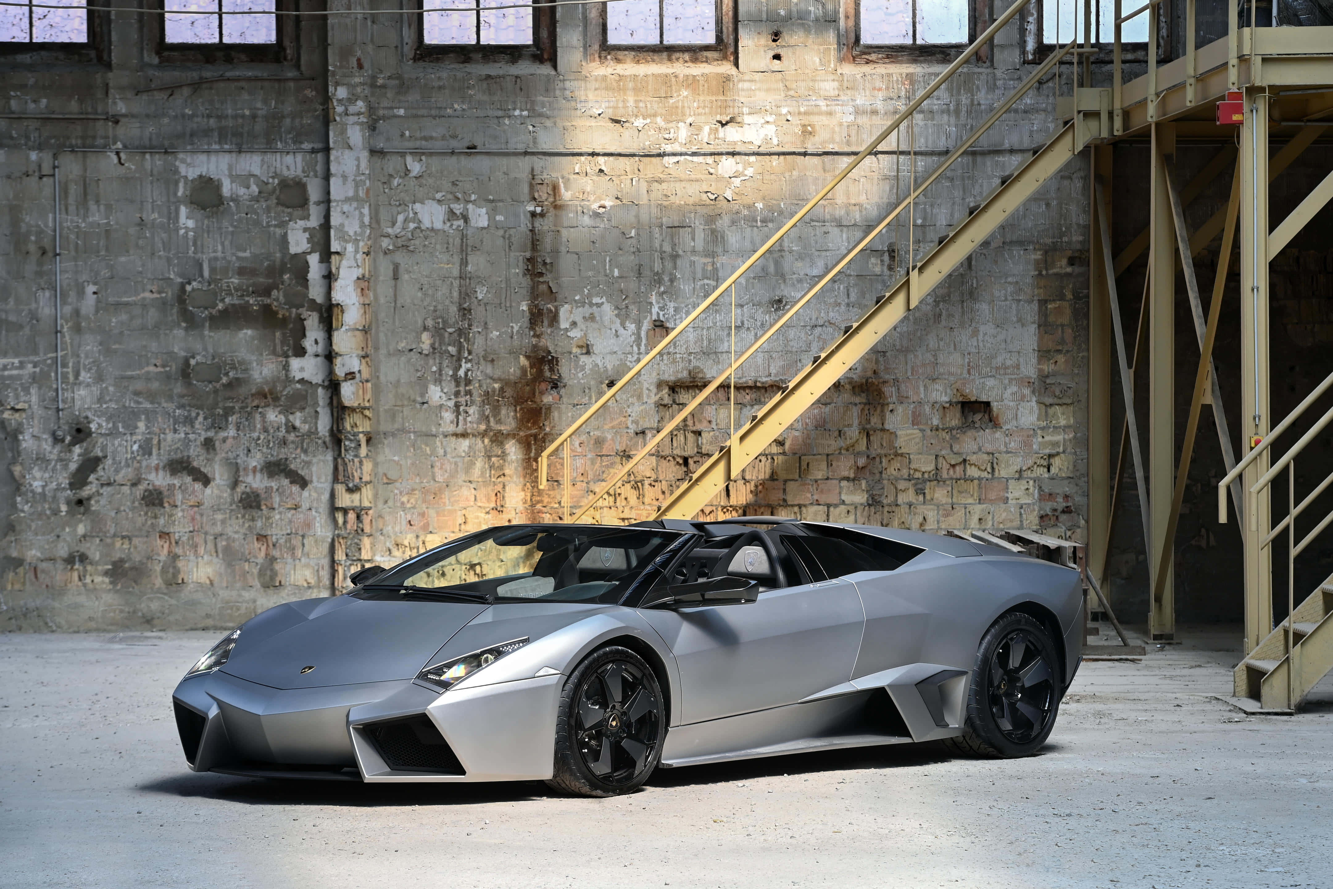 Lamborghini Reventón 桌布 壁紙 — 一台時尚的銀色藍寶堅尼 Reventon Roadster 停放在一個風化的工業倉庫內，倉庫有裸露的混凝土牆和黃色金屬樓梯。