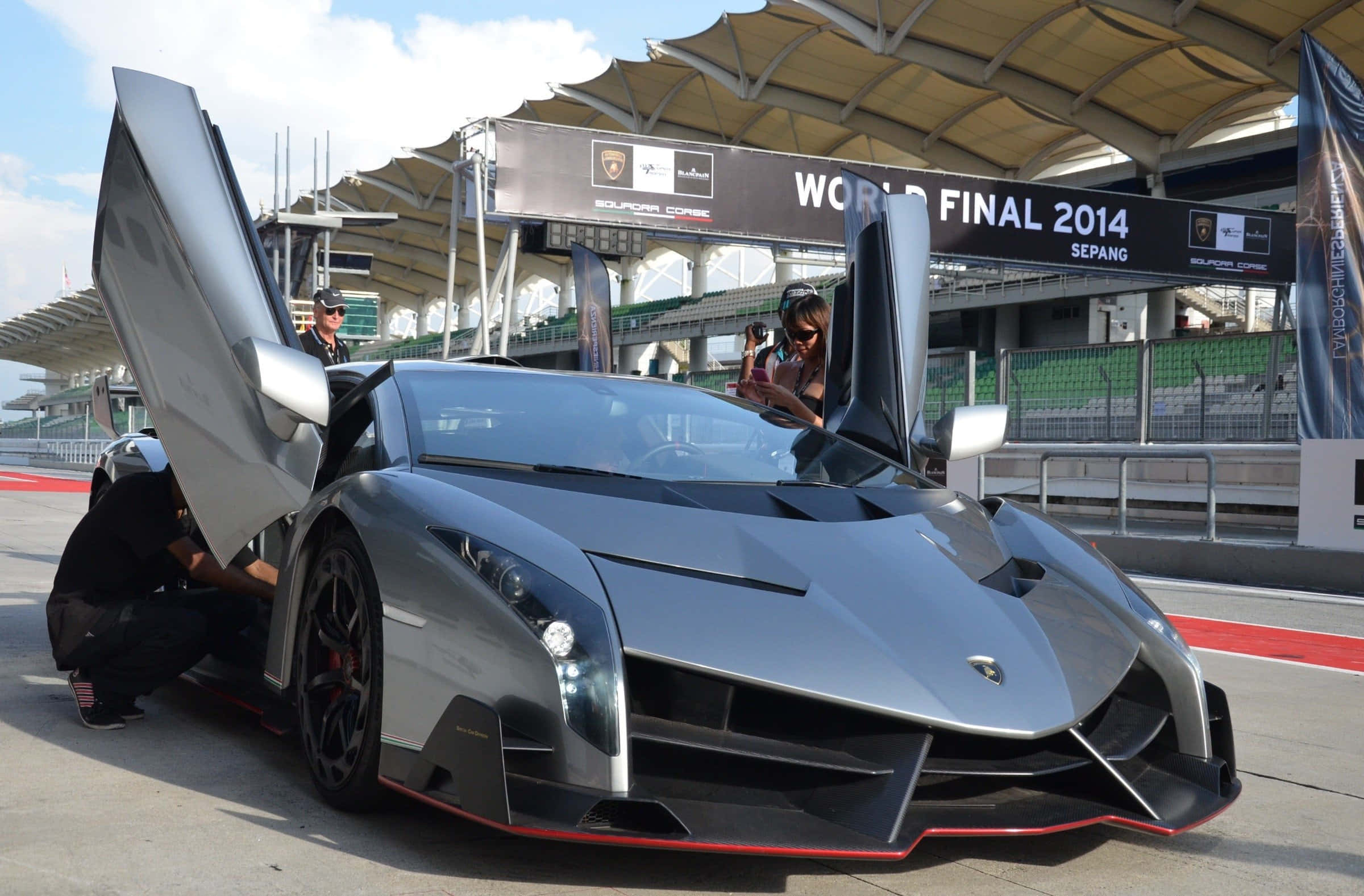 Lamborghini Veneno 桌布 壁紙 — 一架流線型銀色藍寶堅尼 Veneno 在賽道上打開鷗翼門，工作人員在明亮的日光下圍繞它工作。