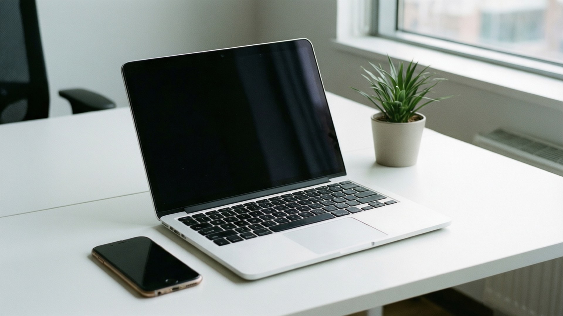 Minimalist Desktop Setup 壁紙 — 一台時尚的銀色筆記型電腦，配備黑色鍵盤，打開放在白色桌子上，旁邊有一部智慧型手機和一個小盆栽多肉植物，靠近窗戶。