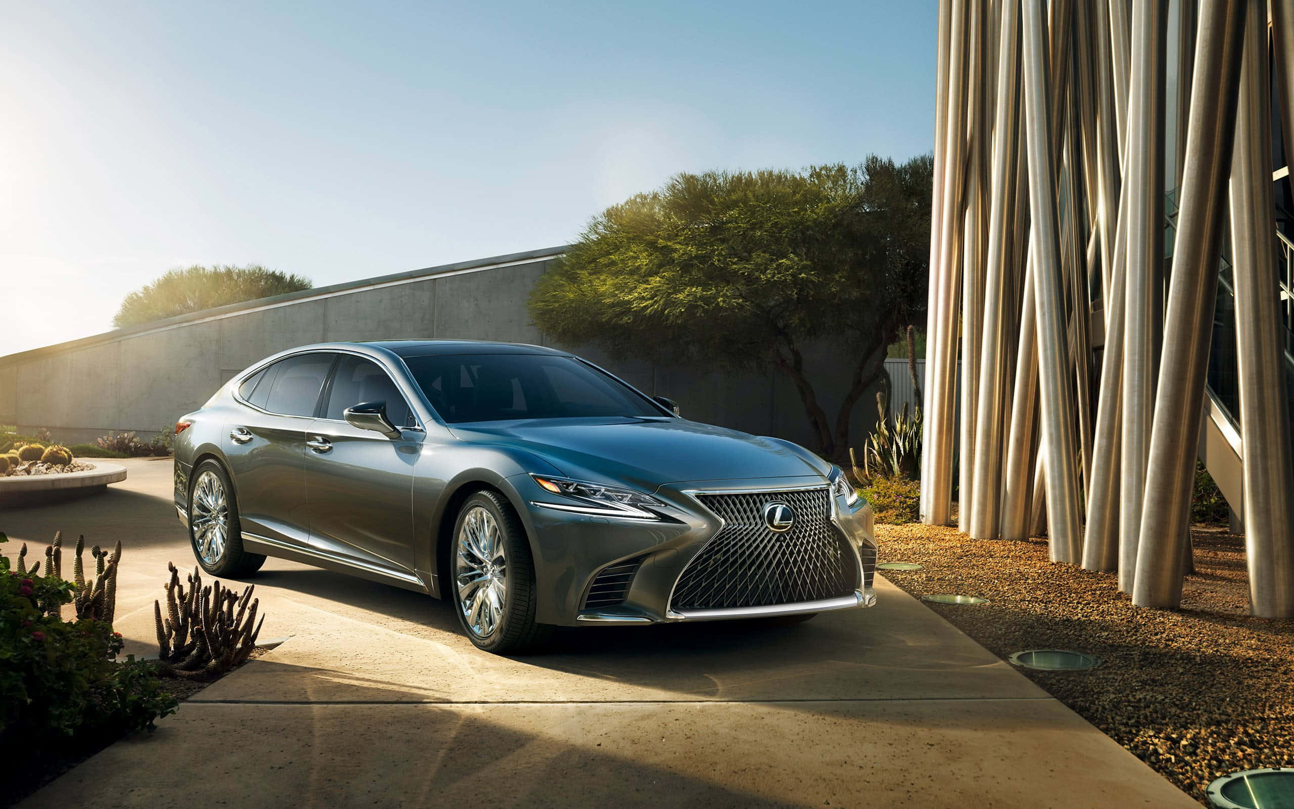 Lexus Ls 500 桌布 壁紙 — 一輛時尚的銀色Lexus轎車停放在陽光普照的混凝土車道上，旁邊是現代建築和沙漠植物。