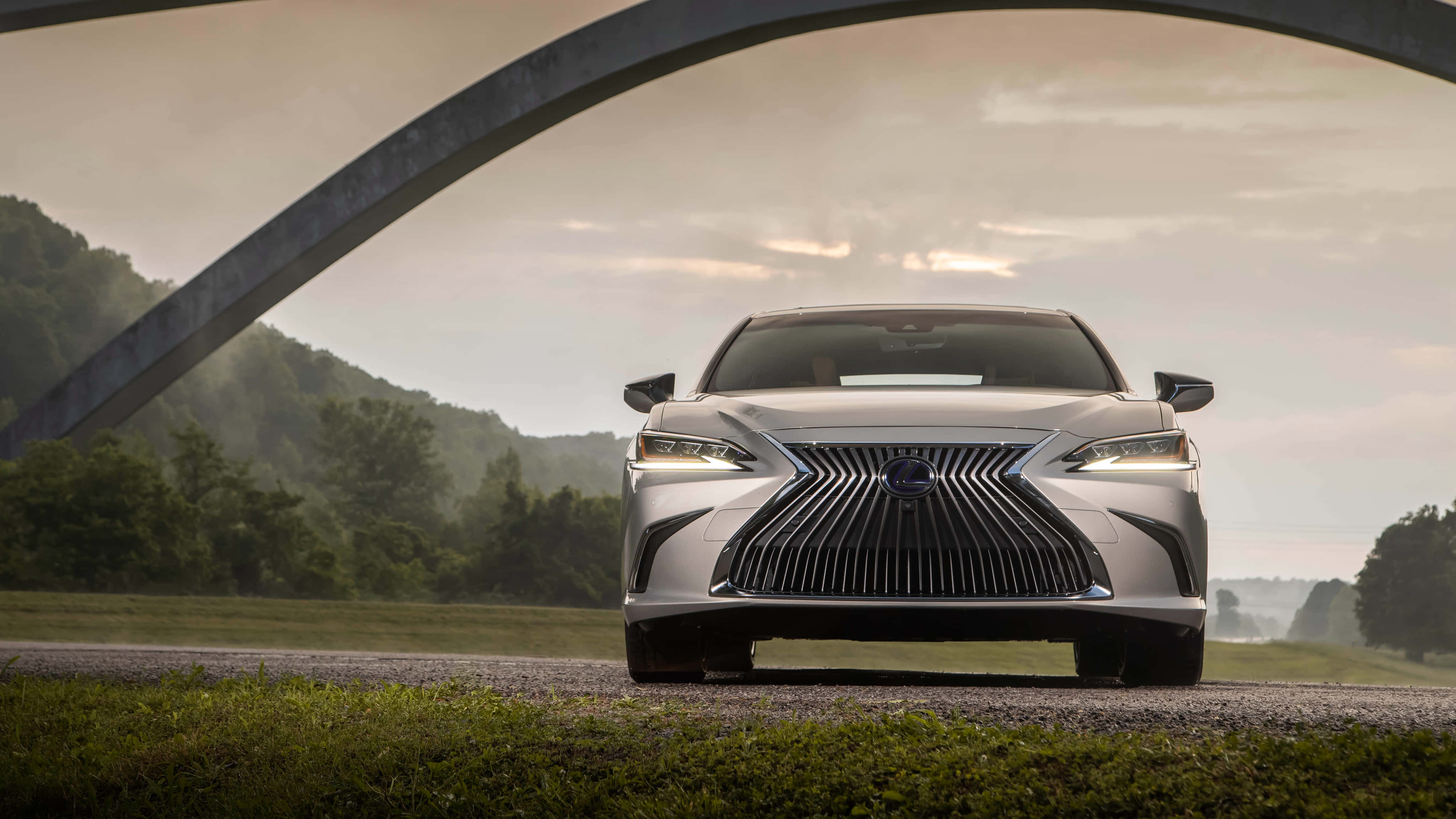 Lexus Es 300h 桌布 壁紙 — 一輛時尚的銀色凌志房車在大型彎曲金屬拱門下向前，背景是森林覆蓋的山丘。