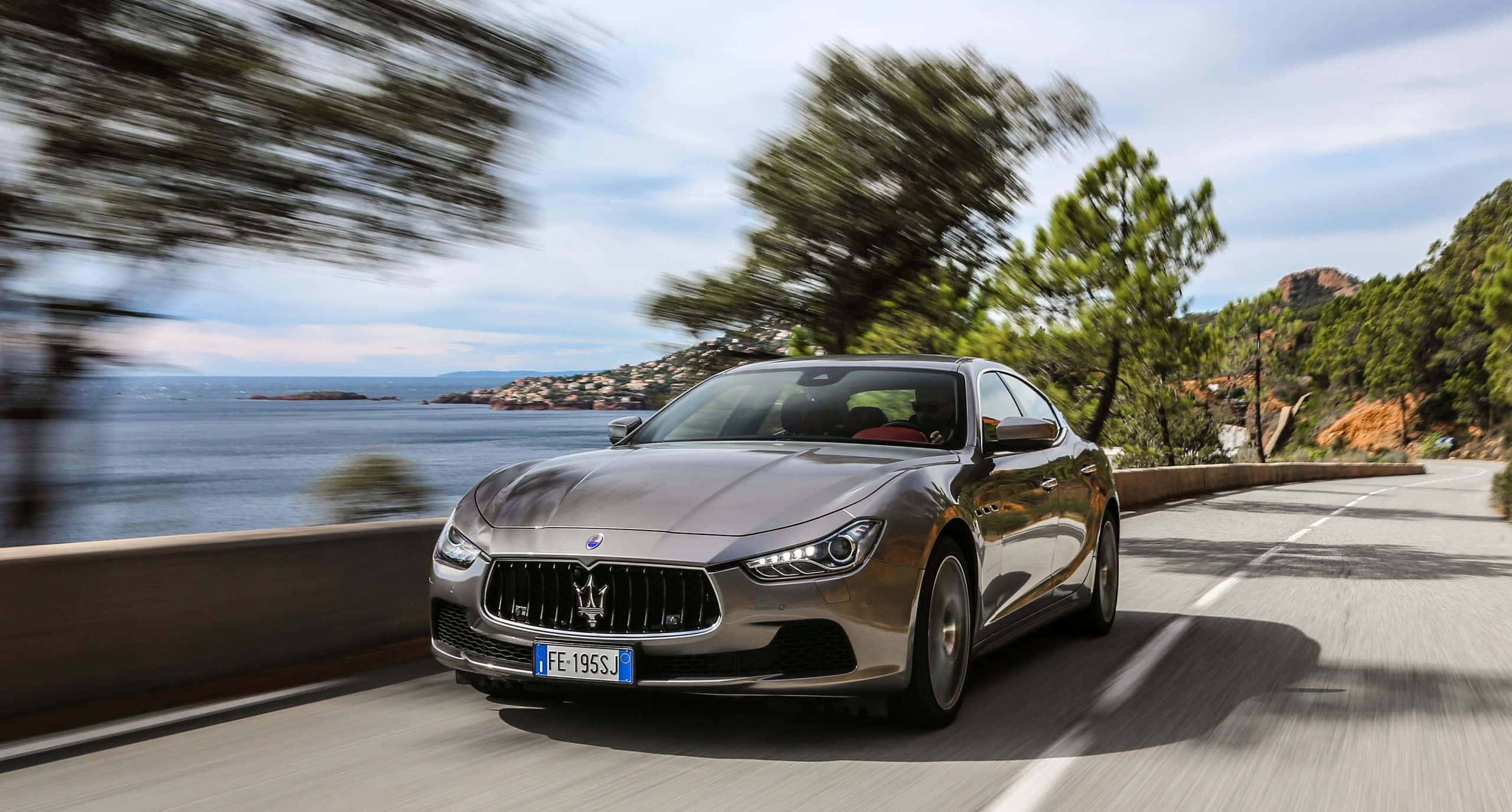 Maserati Ghibli 桌布 壁紙 — 一輛流線型銀色Maserati GranTurismo沿著海岸公路飛馳，背景是模糊的樹木和藍色大海。