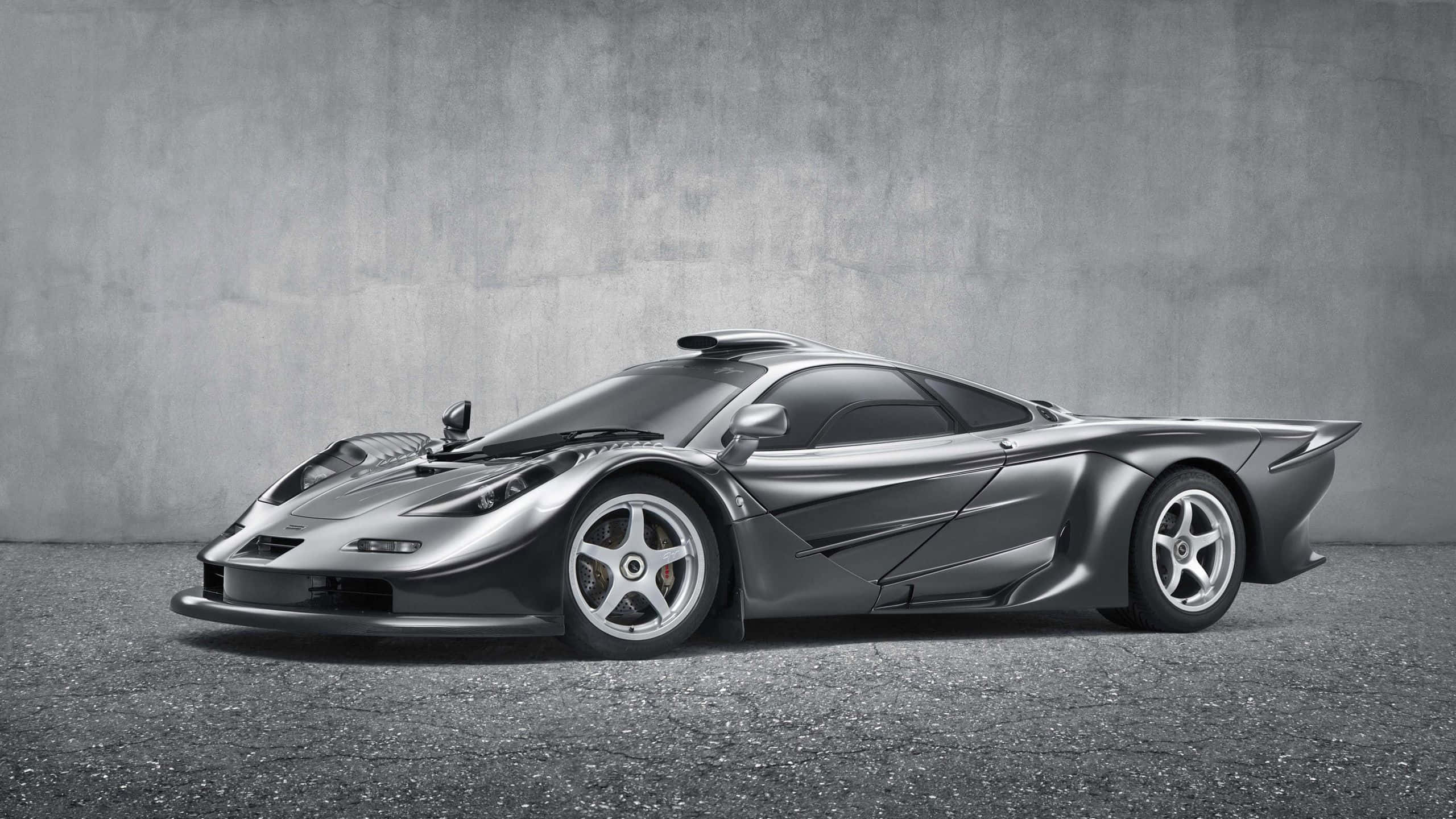 Mclaren F1 桌布 壁紙 — 一輛流線型的銀色McLaren F1超級跑車以側面姿態展示，背景是帶紋理的灰色混凝土牆和瀝青地面。