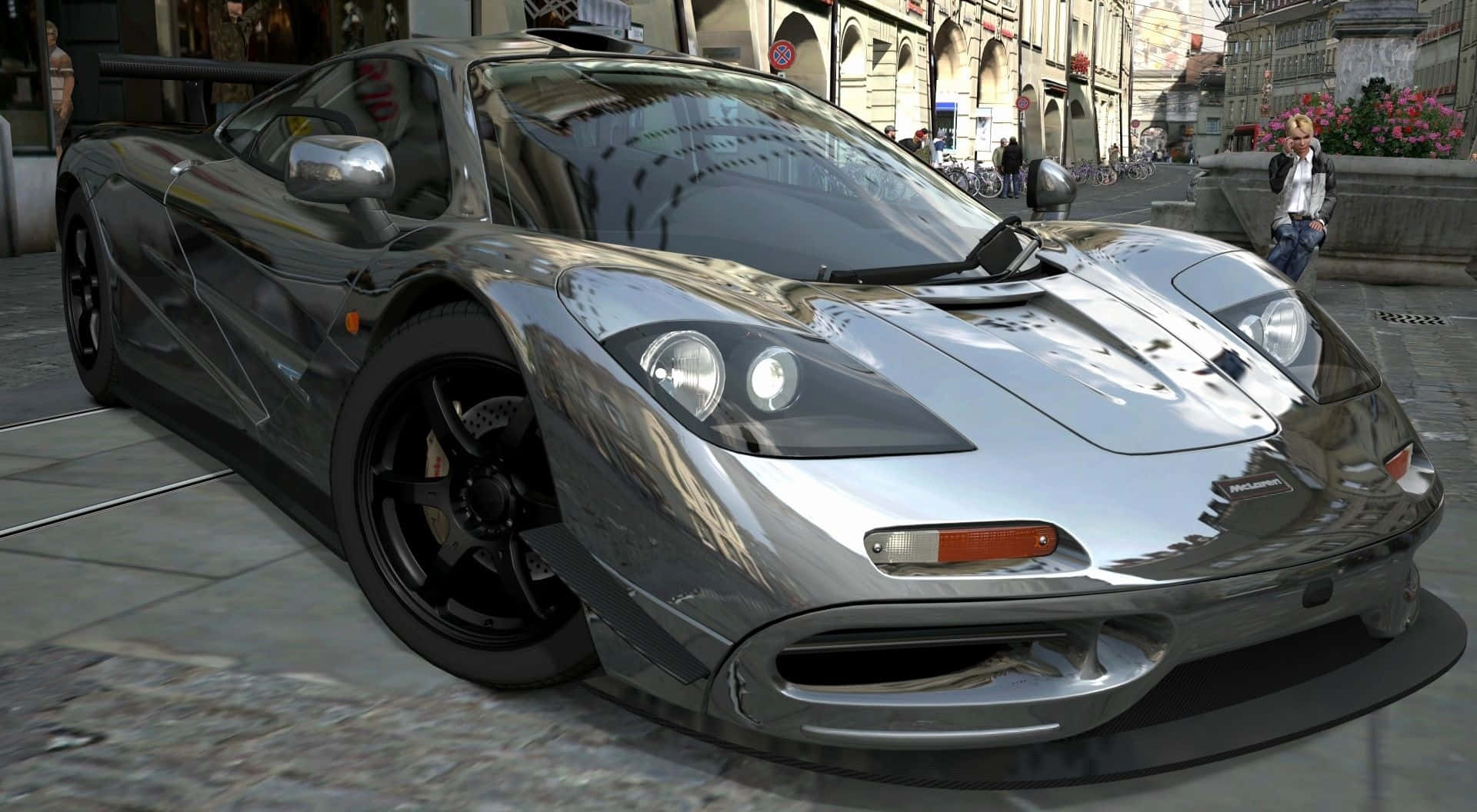 Mclaren F1 桌布 壁紙 — 一輛時尚的銀色McLaren F1超級跑車在歐洲鵝卵石街道上閃耀，周圍有歷史建築和行人，光線柔和。