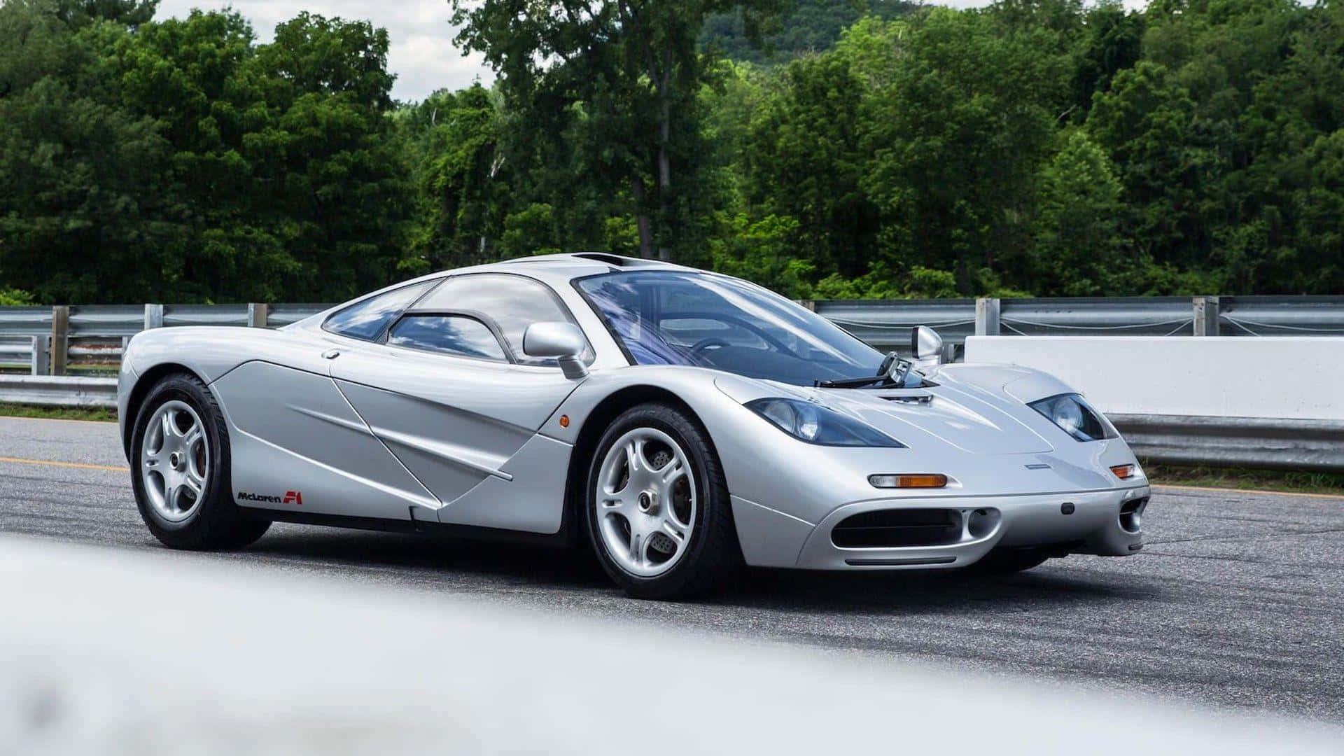 Mclaren F1 桌布 壁紙 — 一輛流線型銀色McLaren F1超級跑車沿著蜿蜒的道路行駛，兩旁是茂密的綠樹和金屬護欄。