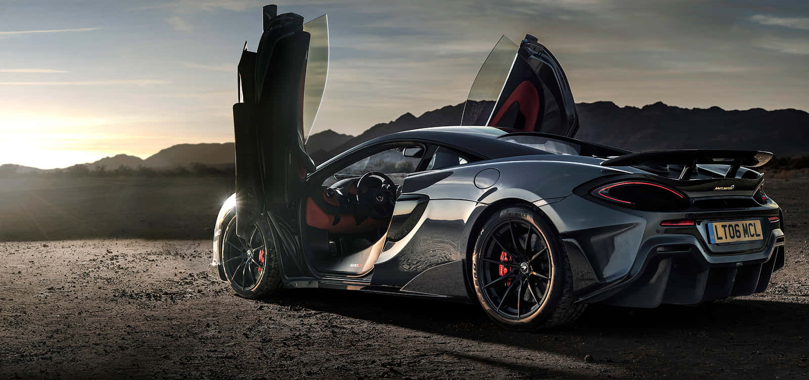 Mclaren 600lt 桌布 壁紙 — 一輛線條流暢的銀色McLaren跑車，鷗翼車門打開，在日落時分的沙漠中，背景有山脈。