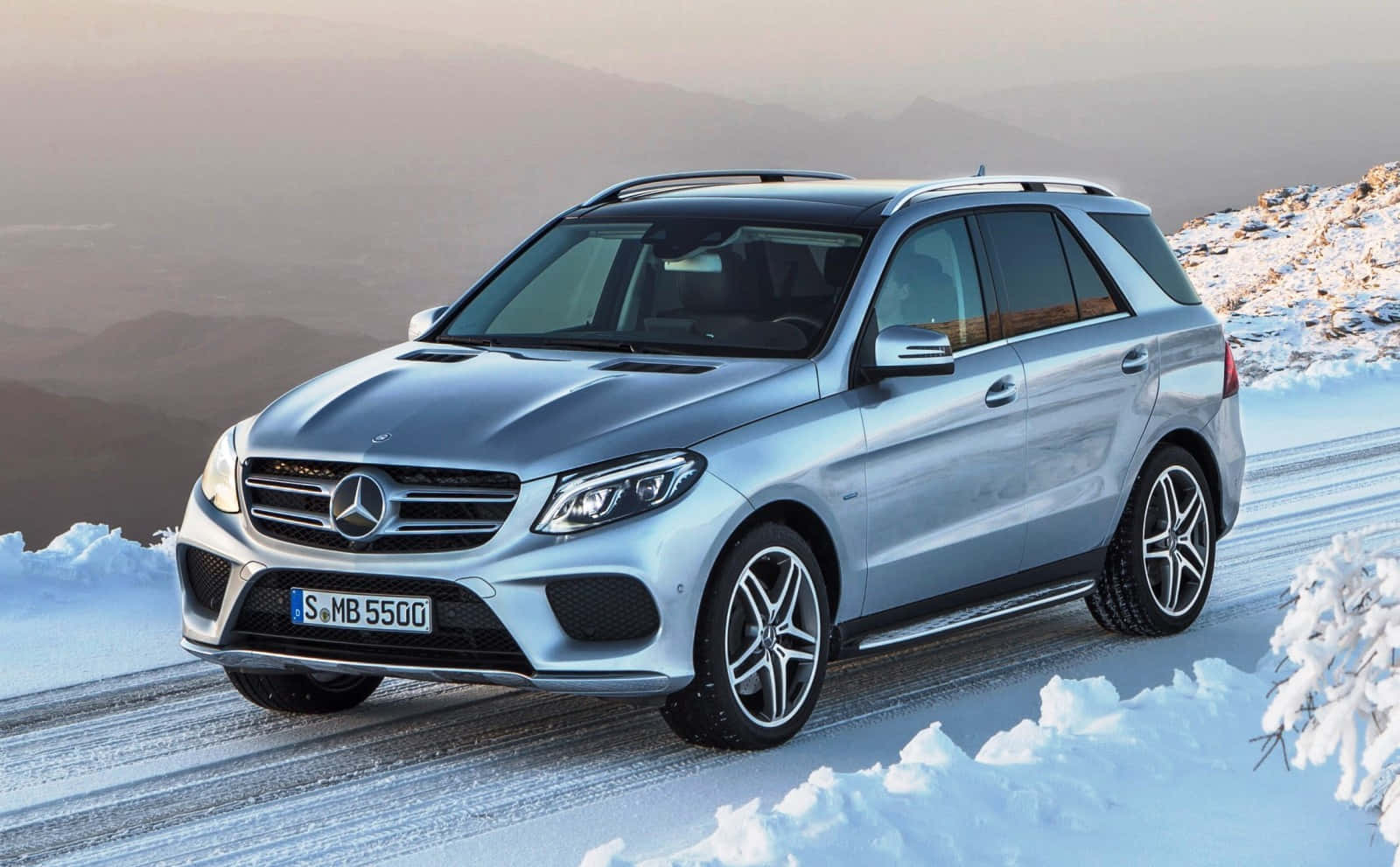 Mercedes Benz Gle-class 桌布 壁紙 — 一輛時尚的銀色梅賽德斯-奔馳SUV在白雪覆蓋的山路上自信行駛，遠方山峰在柔和冬日天空下。