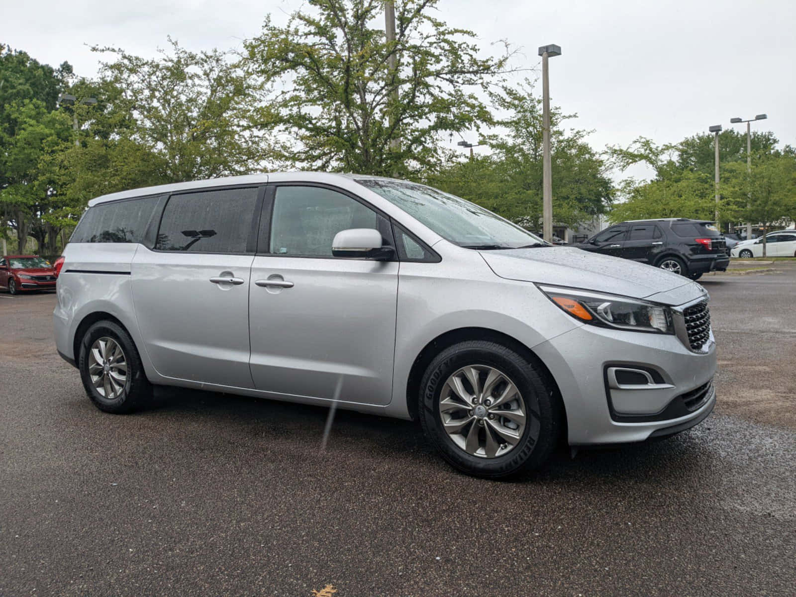 Kia Sedona 桌布 壁紙 — 一輛時尚的銀色小型貨車停放在潮濕的瀝青停車場，周圍綠樹成蔭，天空多雲。