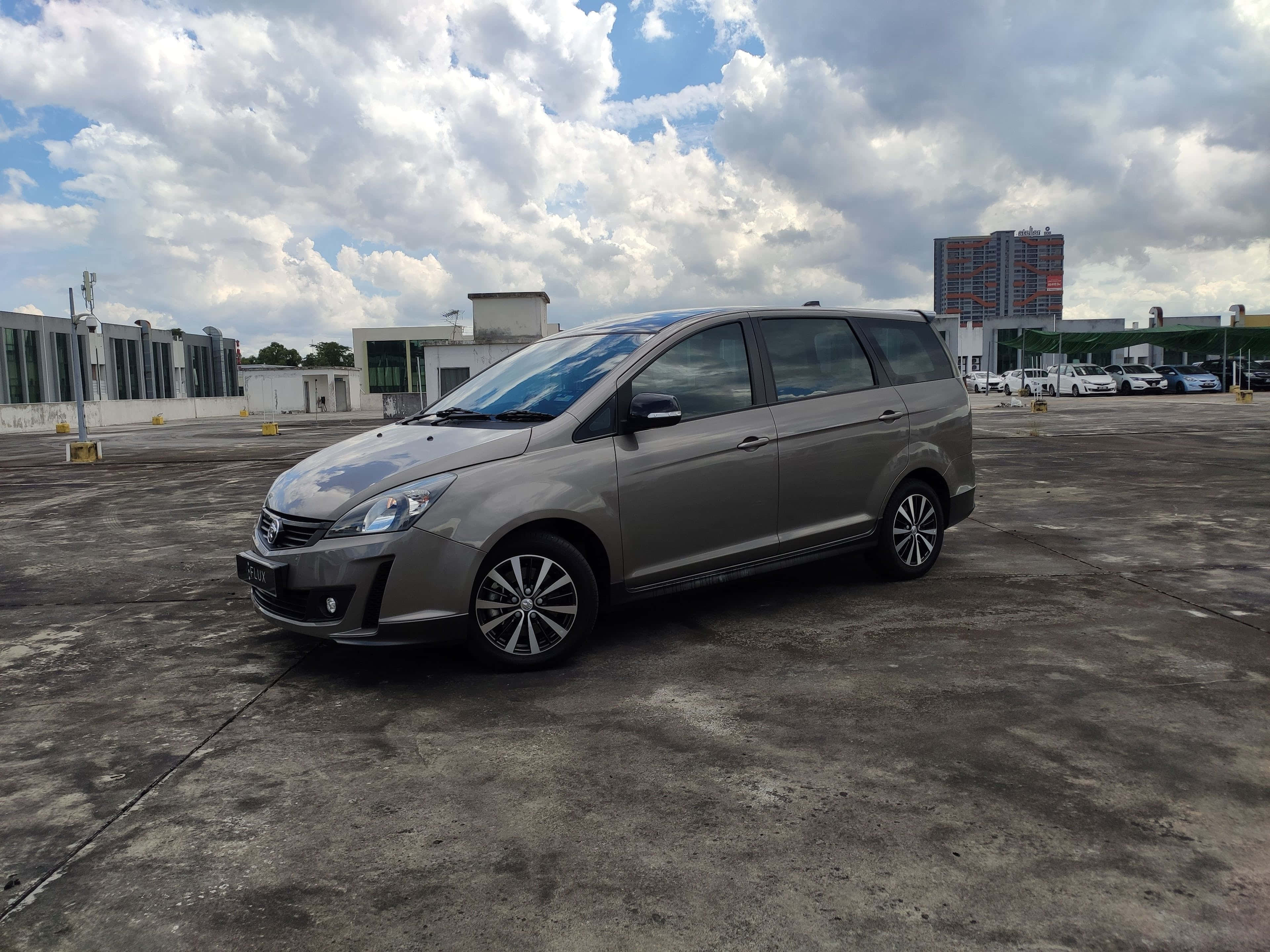Proton Exora 桌布 壁紙 — 一輛時尚的銀色小型貨車停放在廣闊的混凝土場地上，背景是戲劇性的多雲天空，遠處有現代建築。