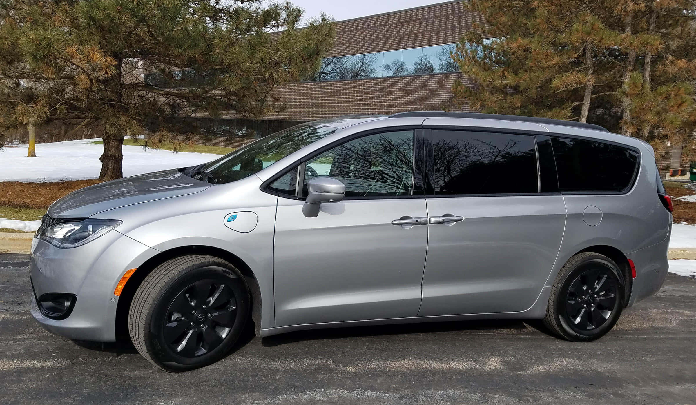 Chrysler Pacifica 桌布 壁紙 — 一輛時尚的銀色小型貨車配黑色輪圈，停放在雪地車道上，旁邊有松樹和現代磚砌建築。