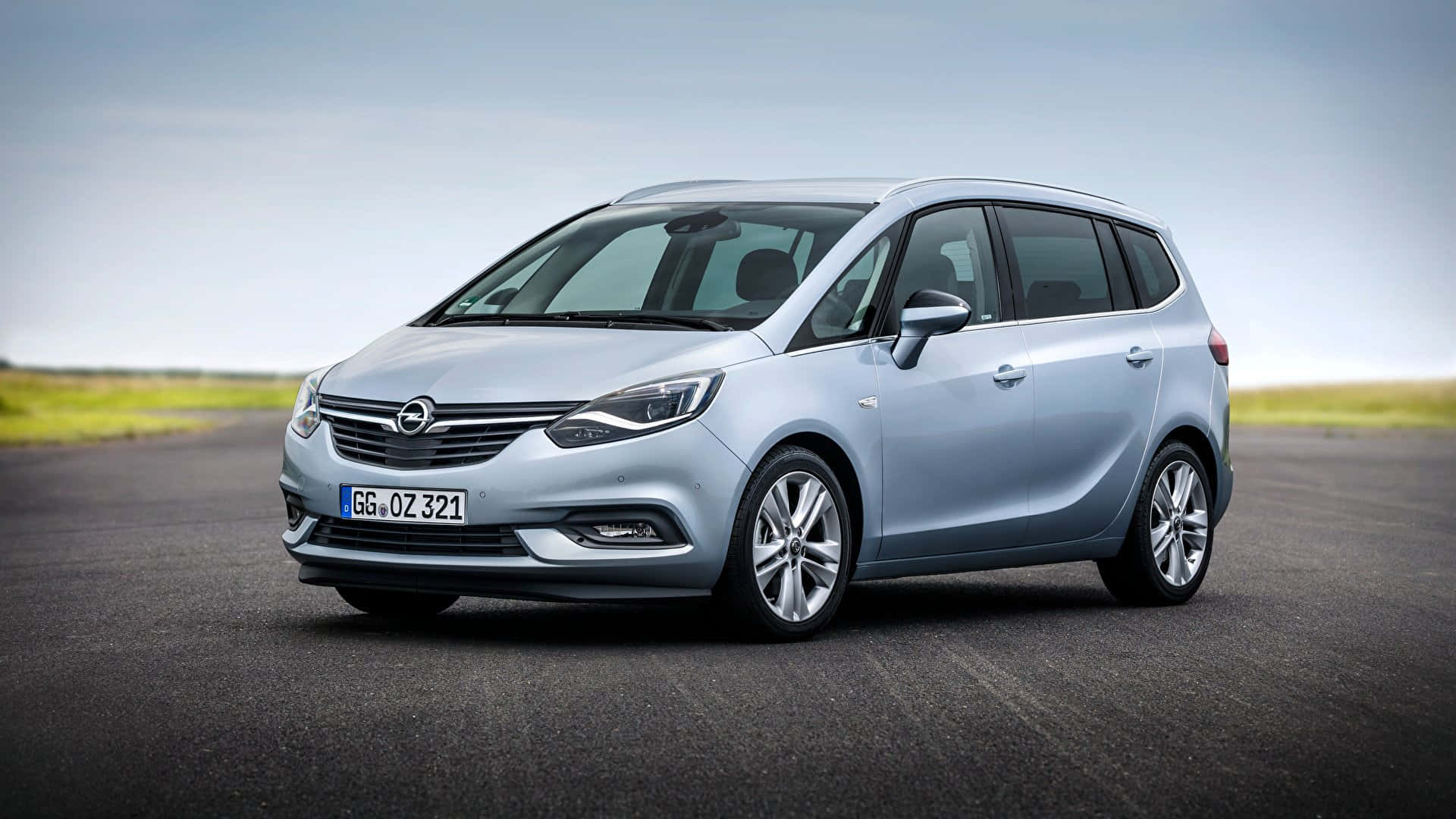 Opel Zafira 桌布 壁紙 — 一輛時尚的銀色歐寶Zafira停放在開闊的柏油路上，旁邊是草地，天空蒼白。