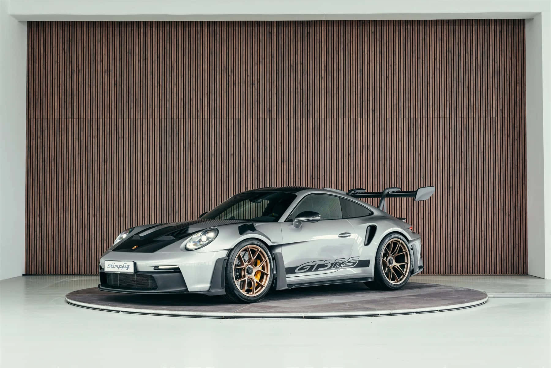  壁紙 — 一輛時尚的銀色保時捷911 GT3 RS配金色輪圈，在極簡主義工作室的木條牆前展示於圓形平台上。