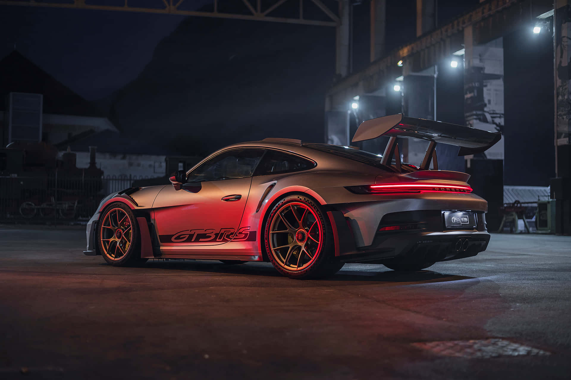  壁紙 — 一輛時尚的銀色保時捷911 GT3 RS，帶有發光的紅色飾件和展開的後尾翼，在夜間工業燈光下矗立。