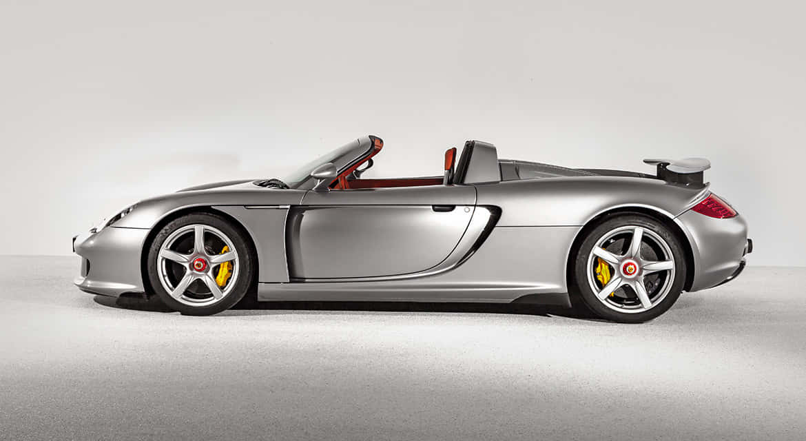 Porsche Carrera Gt 桌布 壁紙 — 一輛流線型銀色保時捷Carrera GT敞篷跑車，搭配紅色內裝及黃色制動卡鉗，佇立於簡約工作室場景中。