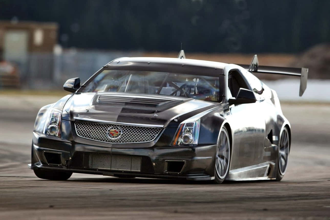 Cadillac Ats 桌布 壁紙 — 一架流線型銀色賽車，帶有大型後翼，在塵土飛揚的賽道上疾馳，背景樹木模糊。