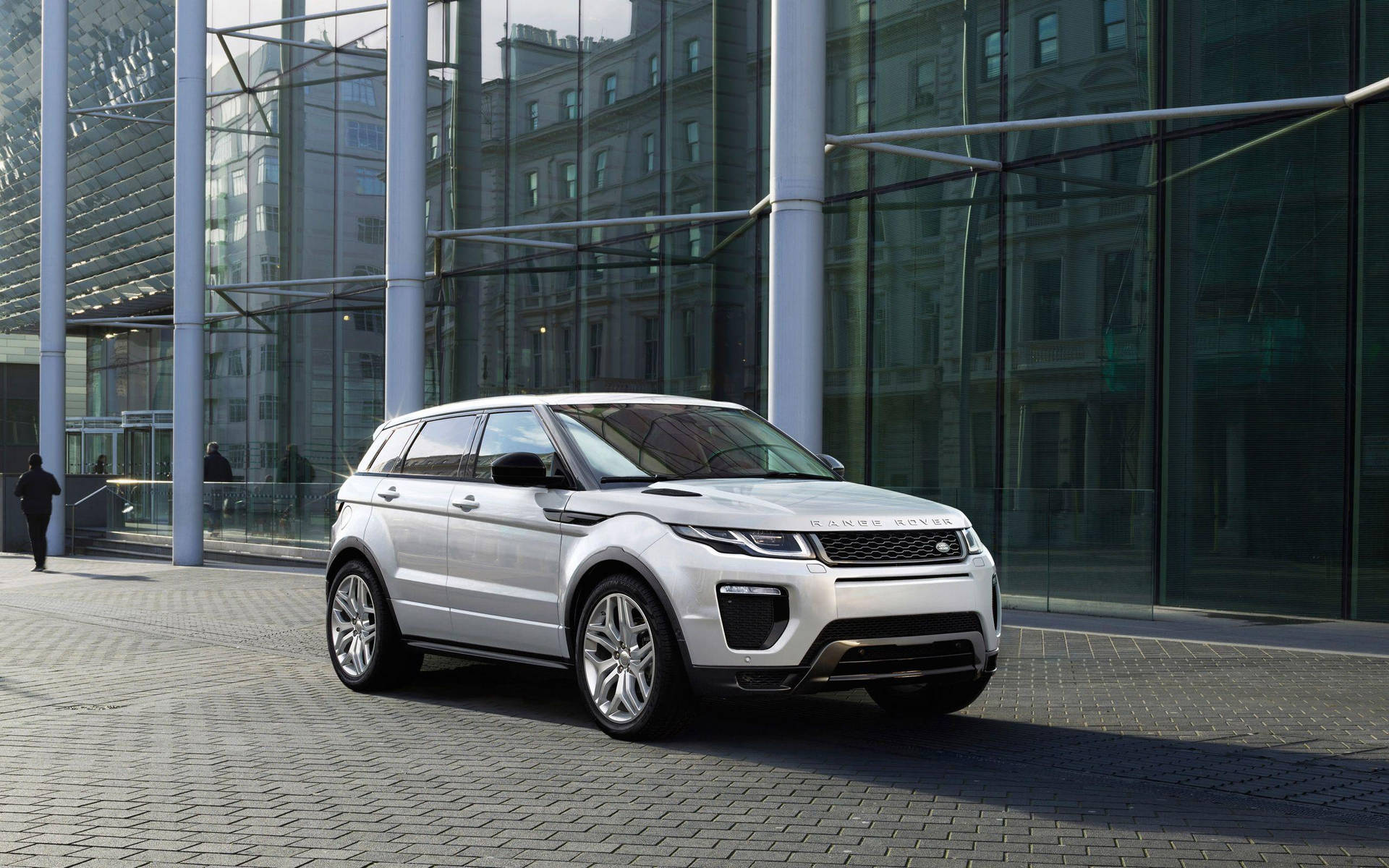 銀色 Range Rover Evoque 停放在玻璃建築前，附近有行人走過。 — 4K 壁紙