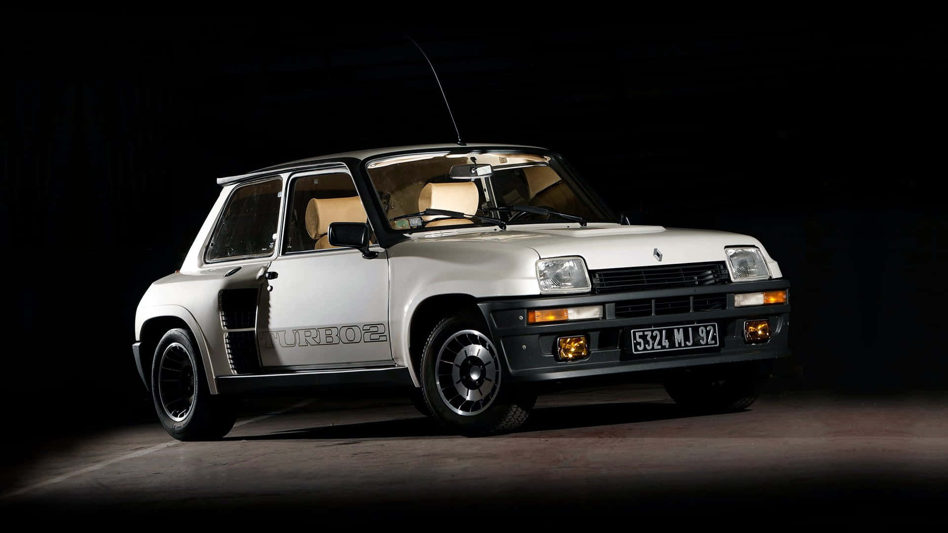 Renault 5 Turbo 桌布 壁紙 — 一輛時尚的銀色雷諾5 Turbo 2跑車在黑色工作室背景前被戲劇性地照亮，聚光燈凸顯其標誌性設計和賽車條紋。