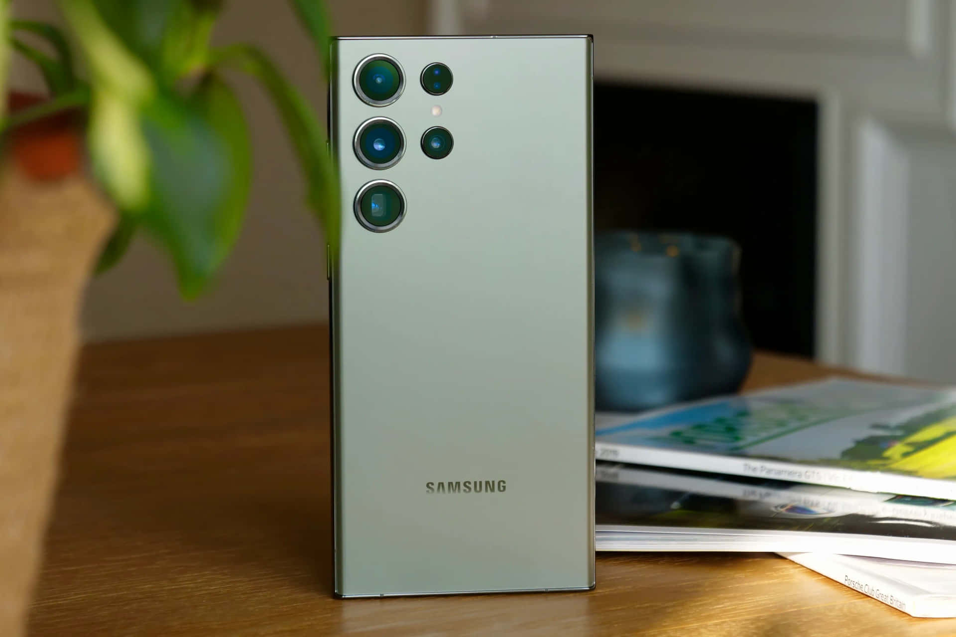  壁紙 — 一部時尚的銀色Samsung智慧型手機直立於木桌上，旁邊有雜誌和盆栽植物。